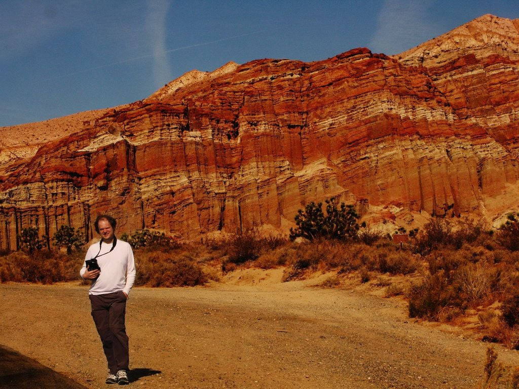 rocky 桌布 壁紙 — 一個人站在泥土路上，面前是湛藍天空下高聳的紅色層狀岩層。