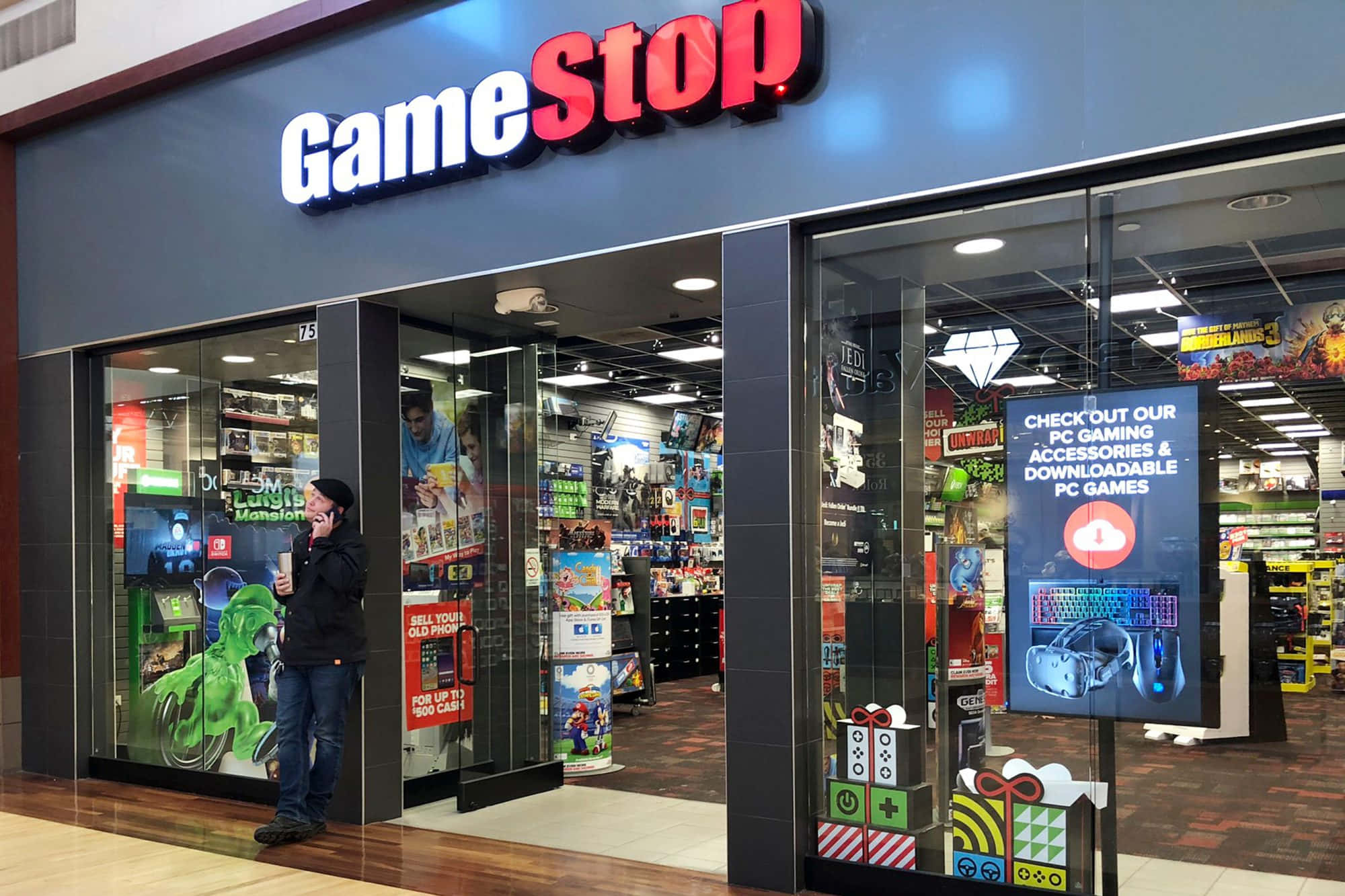 GameStop 桌布 壁紙 — 一個人站在燈光明亮的GameStop商店外，玻璃門內可見色彩繽紛的遊戲商品。