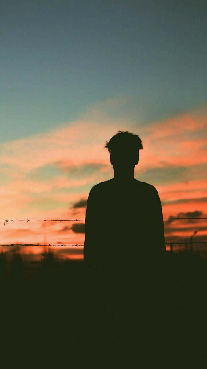 shadow 桌布 壁紙 — 一個人站在鐵絲網後面，背景是充滿橙色和藍色色調的 vibrant 日落天空的剪影。