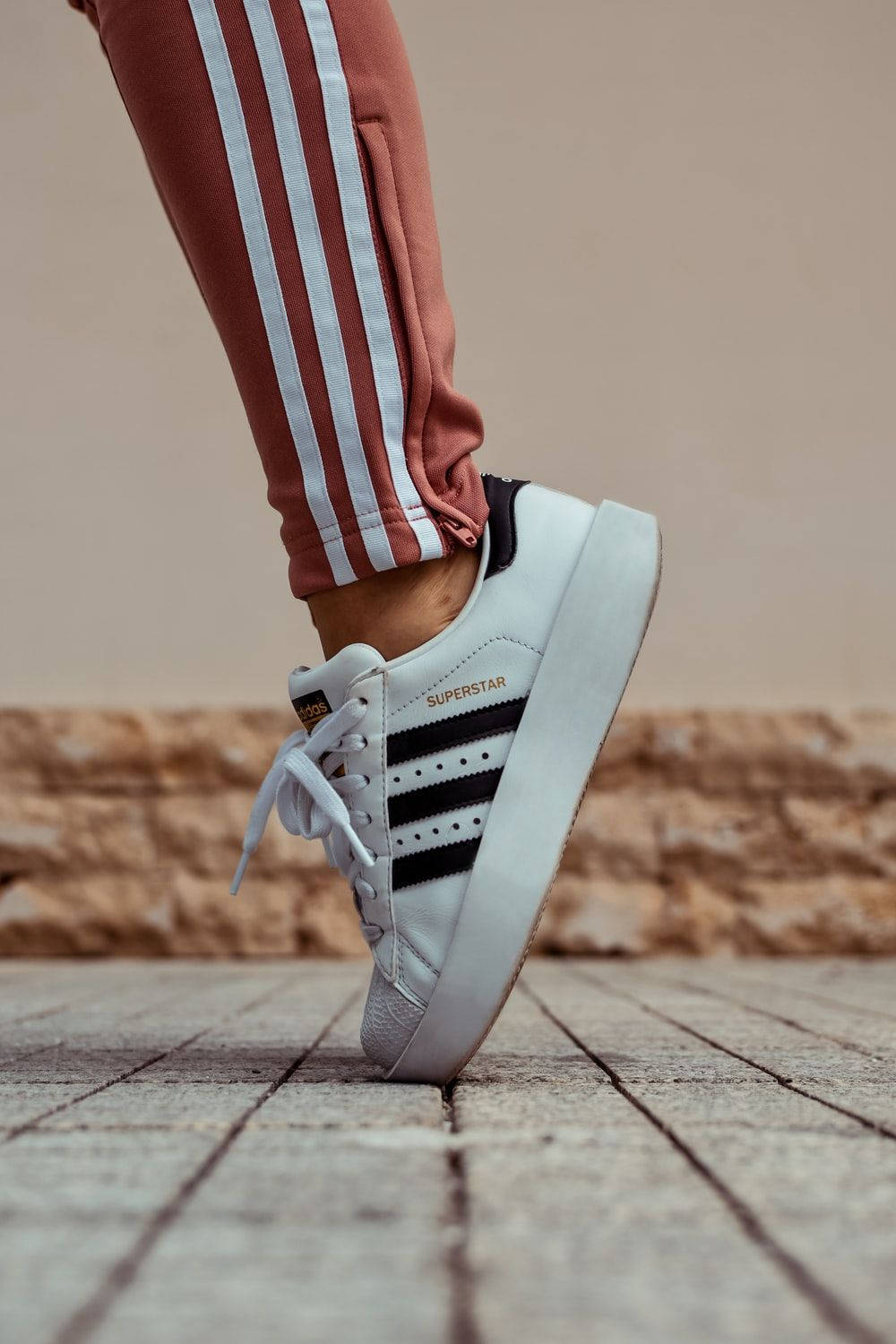 鞋子 桌布 壁紙 — 一個人腳穿白色 adidas superstar 運動鞋，踩在石頭路面上，搭配鏽色運動褲的特寫。