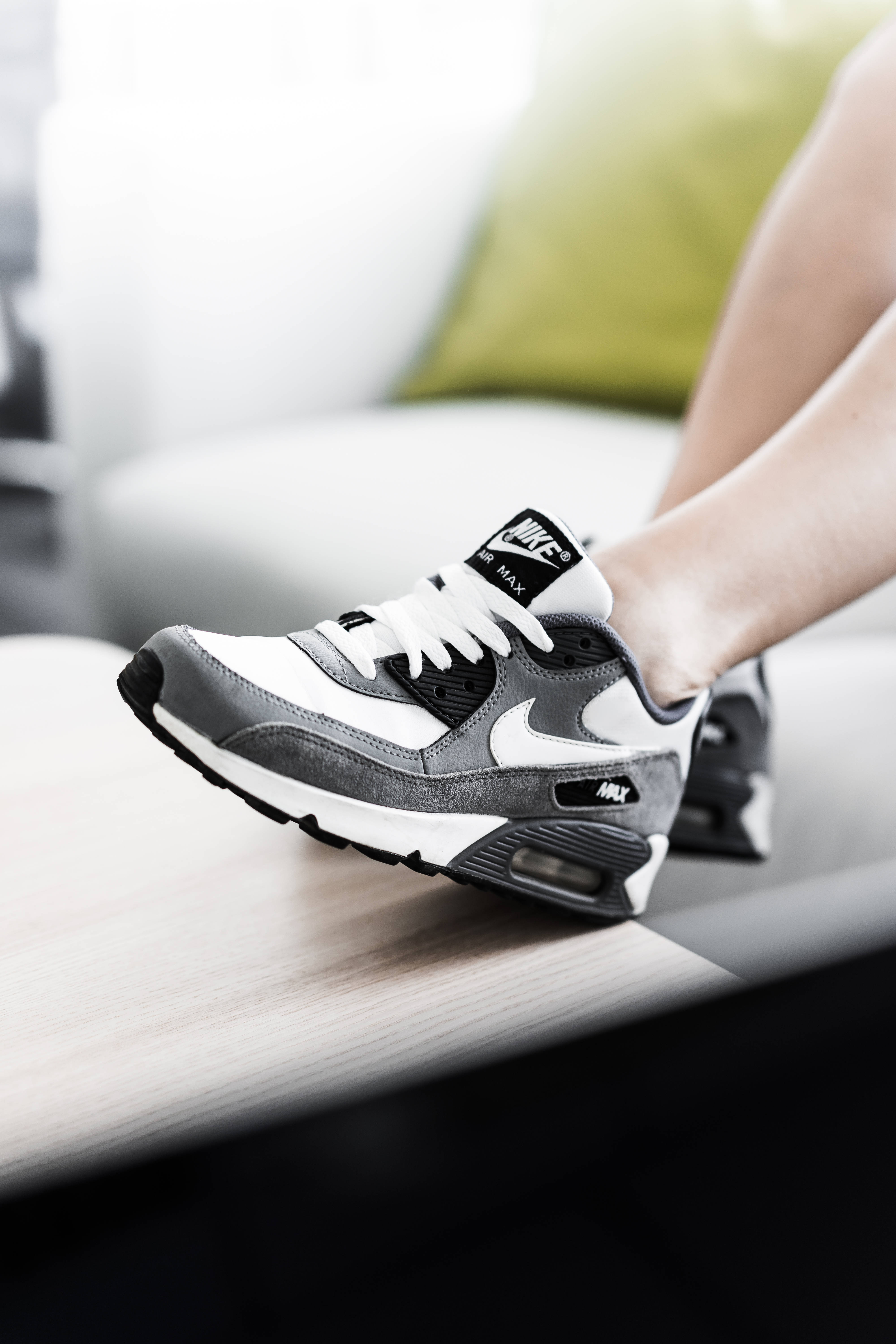 Shoe 桌布 壁紙 — 一個人腳部穿著灰色和白色Nike Air Max運動鞋的特寫，鞋放在木質表面上，背景是模糊的綠色軟墊。