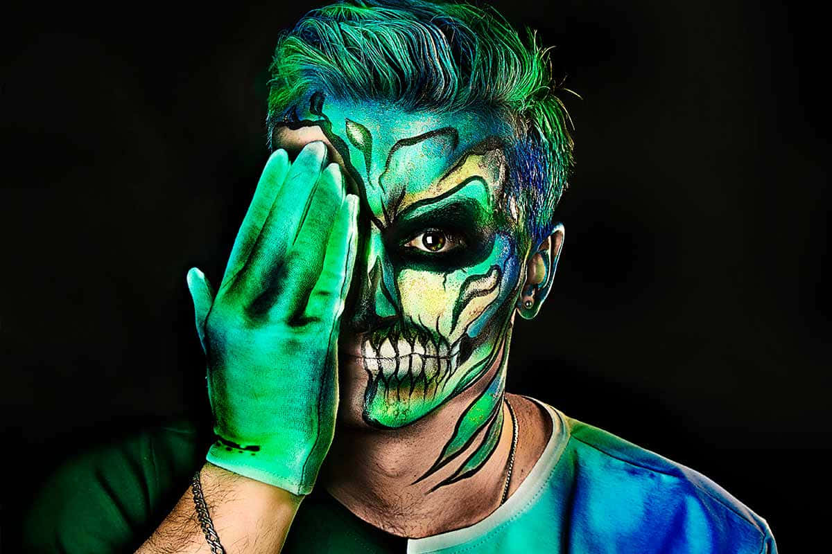 Halloween Face Paint 桌布 壁紙 — 一個人臉上塗有發光的綠色骷髏妝，用戴著相配手套的手遮住半張臉，背景為黑色。