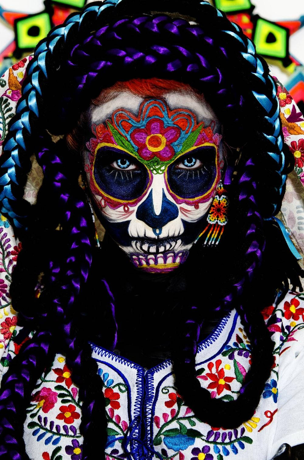 Dia De Los Muertos 桌布 壁紙 — 一個人臉上畫著鮮豔的糖骷髏妝容，梳著色彩繽紛的辮子，穿著花卉刺繡服裝，背景是明亮的圖案。