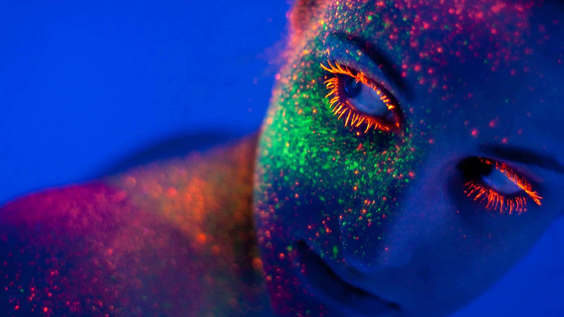 glow 桌布 壁紙 — 一個人臉部的特寫，在黑光下覆蓋著發光的霓虹閃粉，帶有鮮豔的藍色、綠色和橙色色調。