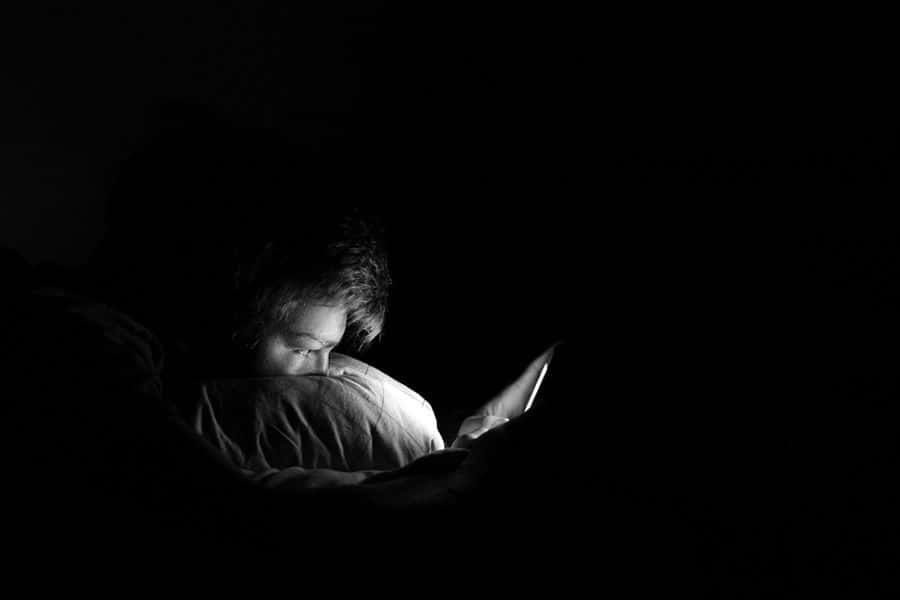 emotion 桌布 壁紙 — 一個人蜷縮在床上被窩中，被黑暗房間中手持裝置的光芒照亮。