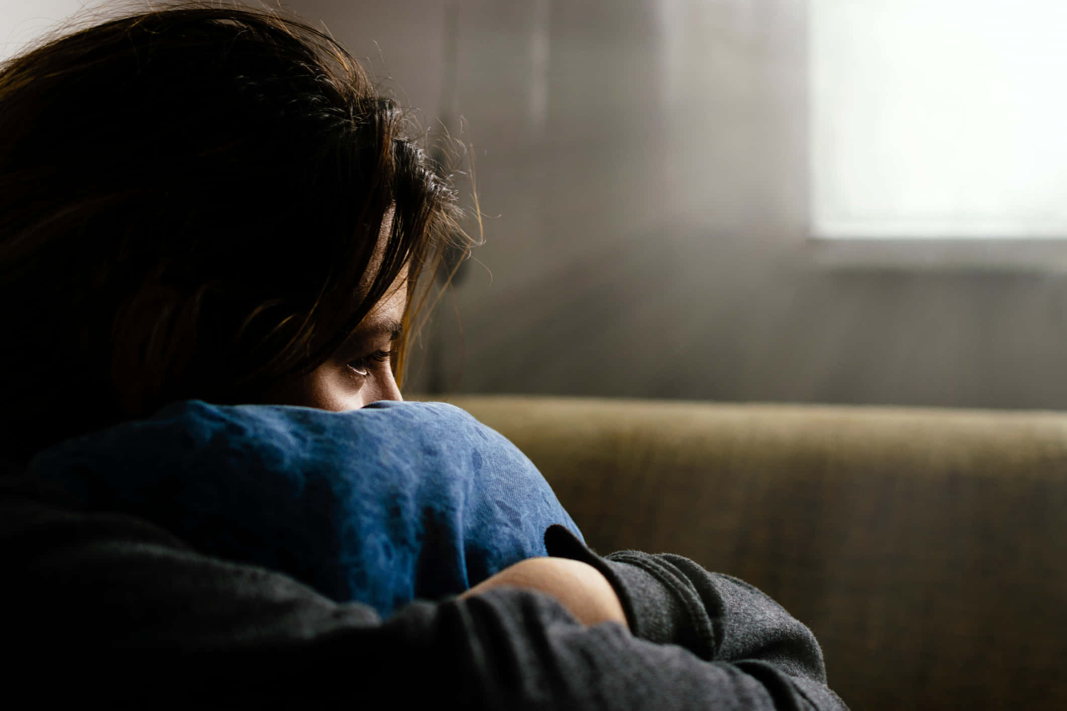Depression Girl 桌布 壁紙 — 一個人蜷縮在沙發上，臉部部分被藍色枕頭遮住，沐浴在柔和窗光中。