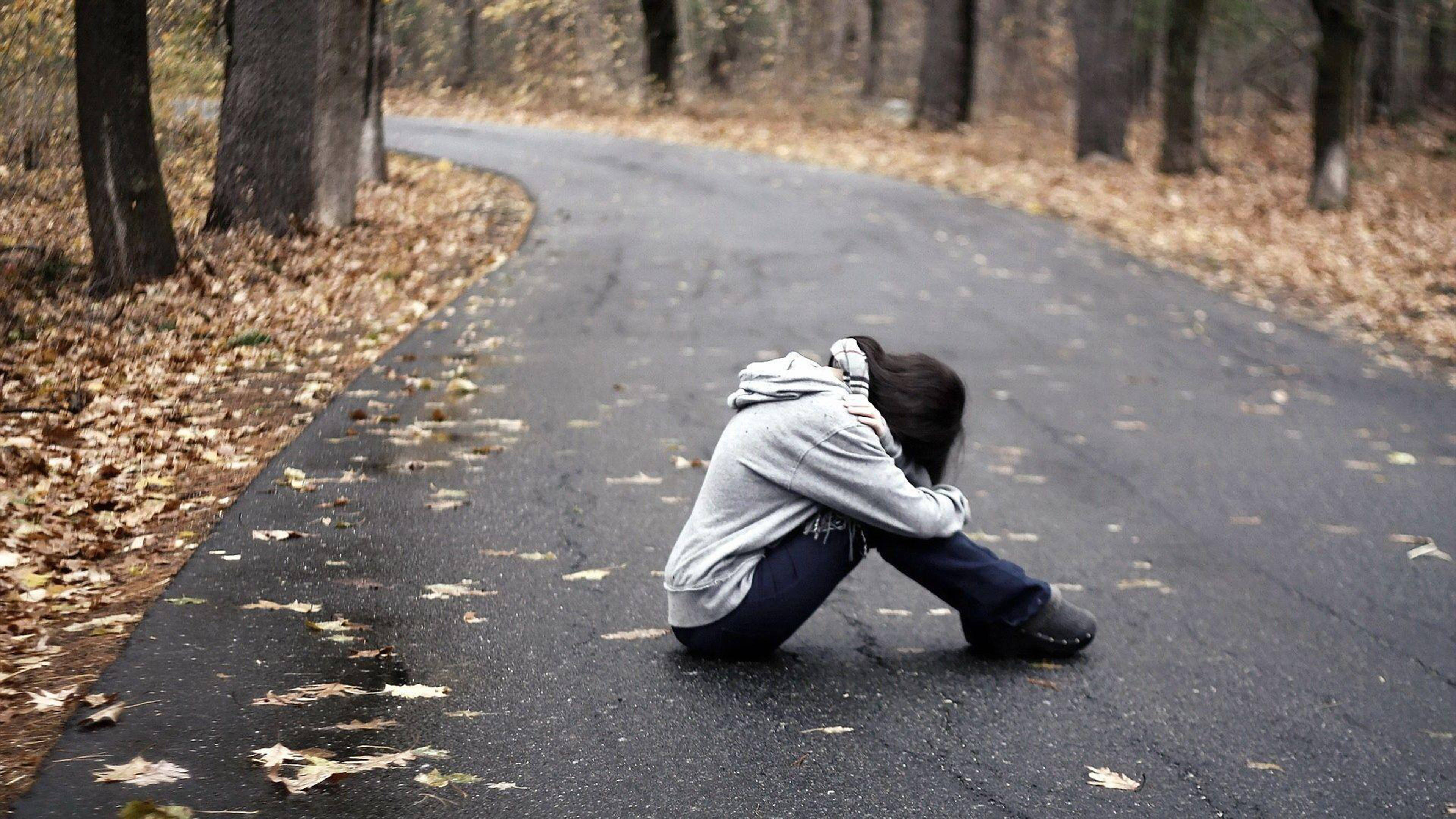 心碎 桌布 壁紙 — 一個人蜷縮在濕漉漉的森林道路上，周圍是秋天的落葉和光禿禿的樹木。