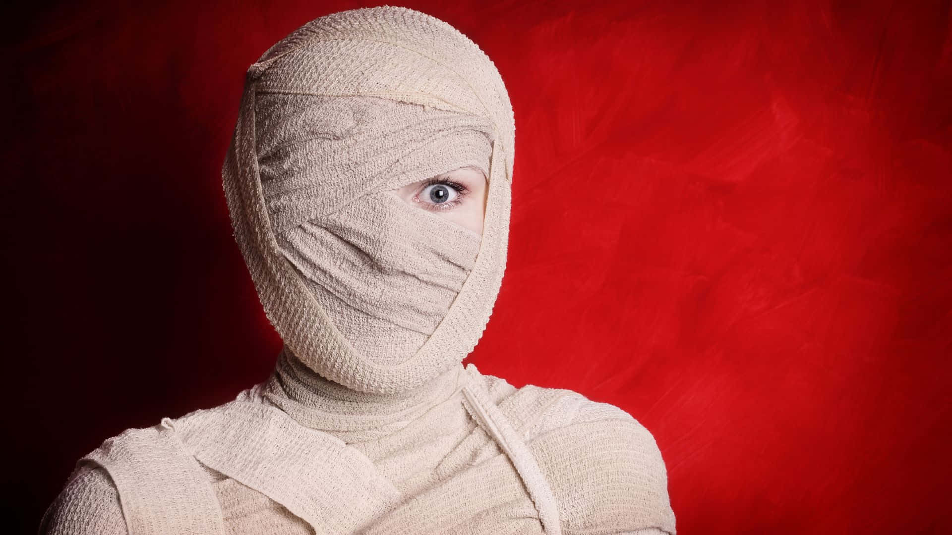 Halloween Costumes 桌布 壁紙 — 一個人被米色繃帶包裹，在紋理紅色背景上露出一隻銳利的藍色眼睛。
