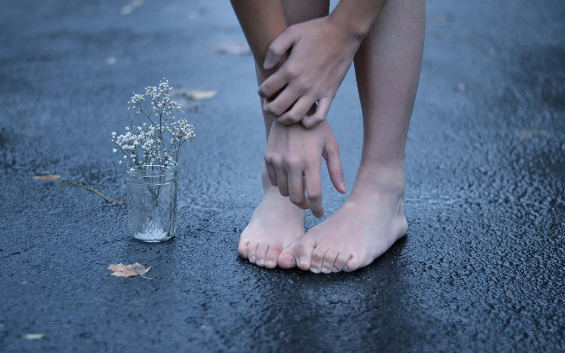 feet 桌布 壁紙 — 一個人赤腳坐在濕漉漉的人行道上，握著腳踝，旁邊有一個裝滿滿天星的小玻璃罐。