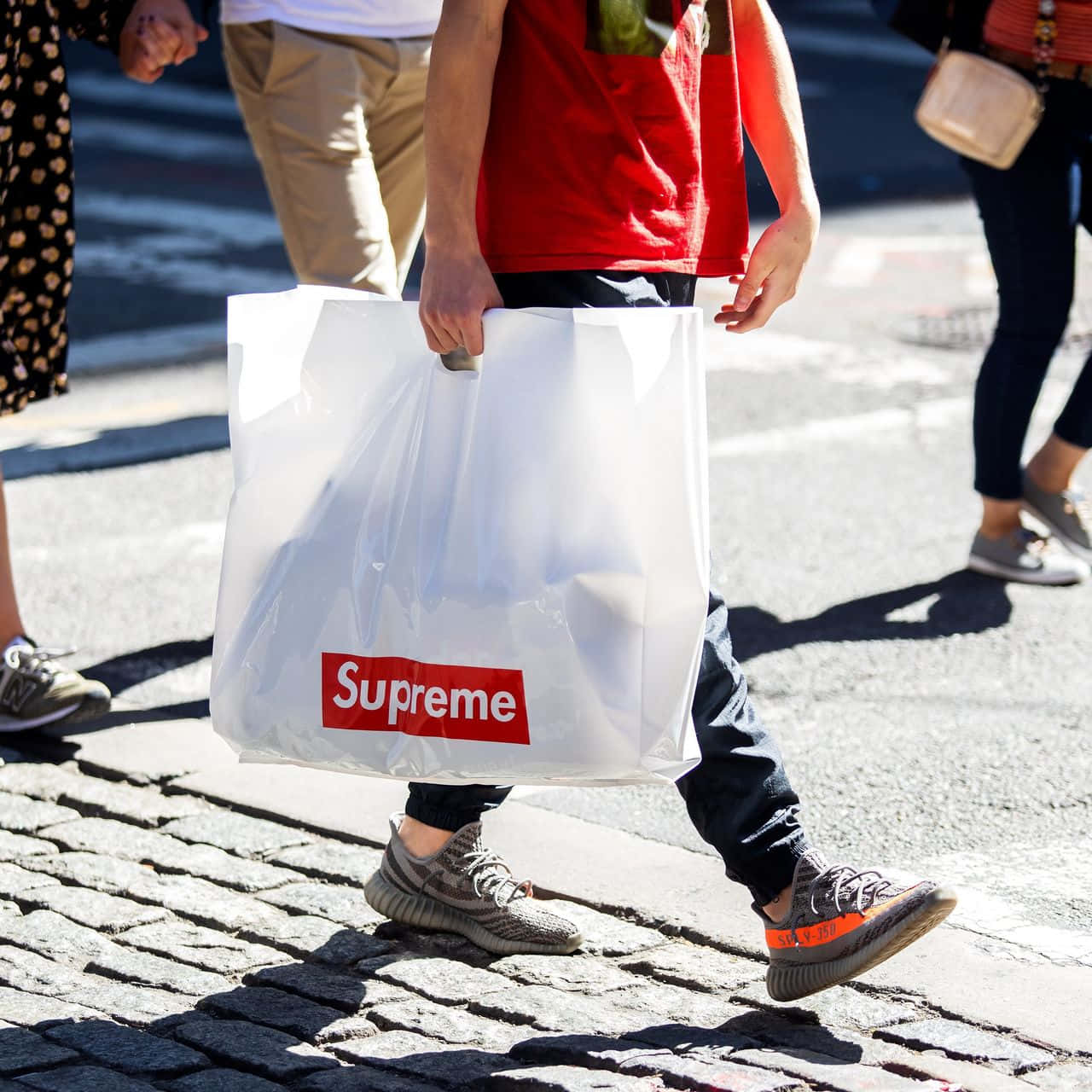 supreme 桌布 壁紙 — 一個人走在鵝卵石上，拿著一個透明的 Supreme 購物袋，上面有粗體的紅色標誌。
