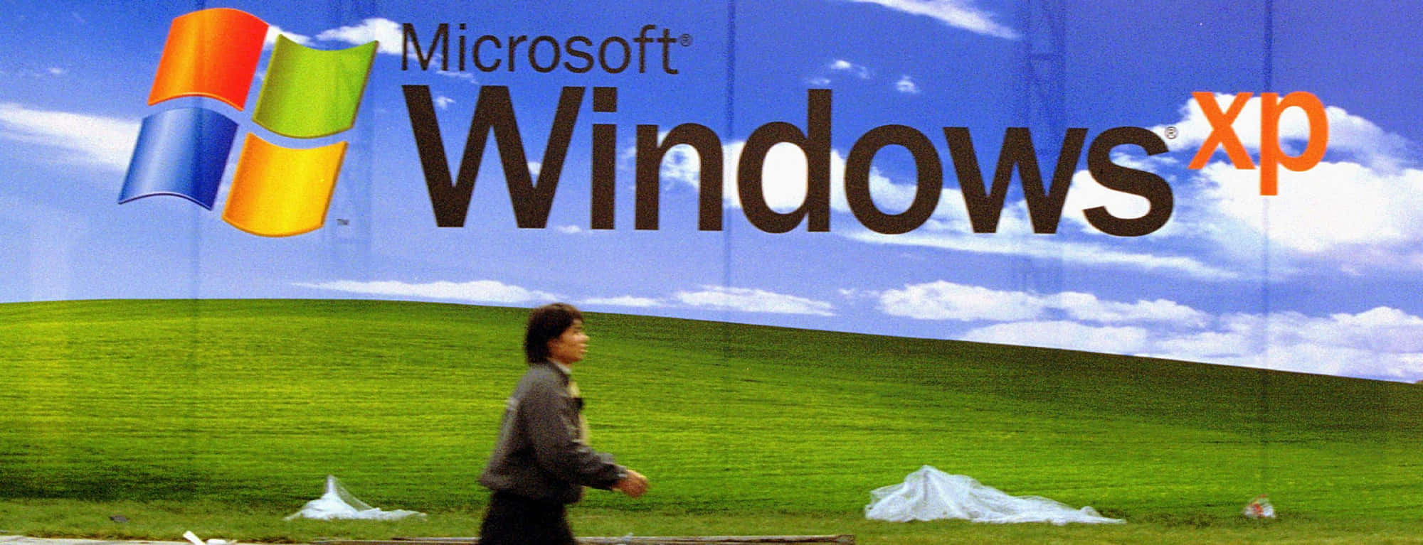 Windows Xp Logo 桌布 壁紙 — 一個人走過一個巨大的廣告牌，上面顯示著標誌性的 Microsoft Windows XP 標誌，背景是綠色山丘和藍天。