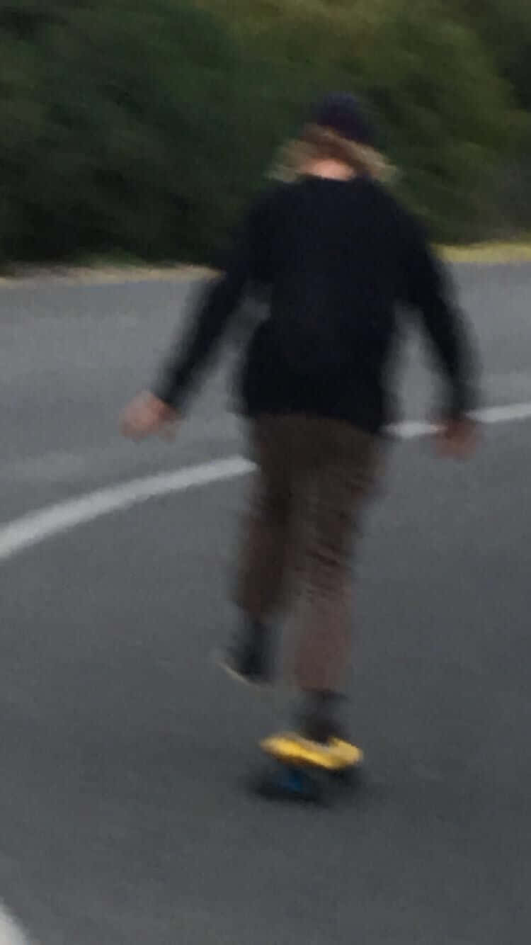 滑板男孩風格 桌布 壁紙 — 一個人踩著滑板沿著彎曲的道路滑下，背景有模糊的綠色植物。