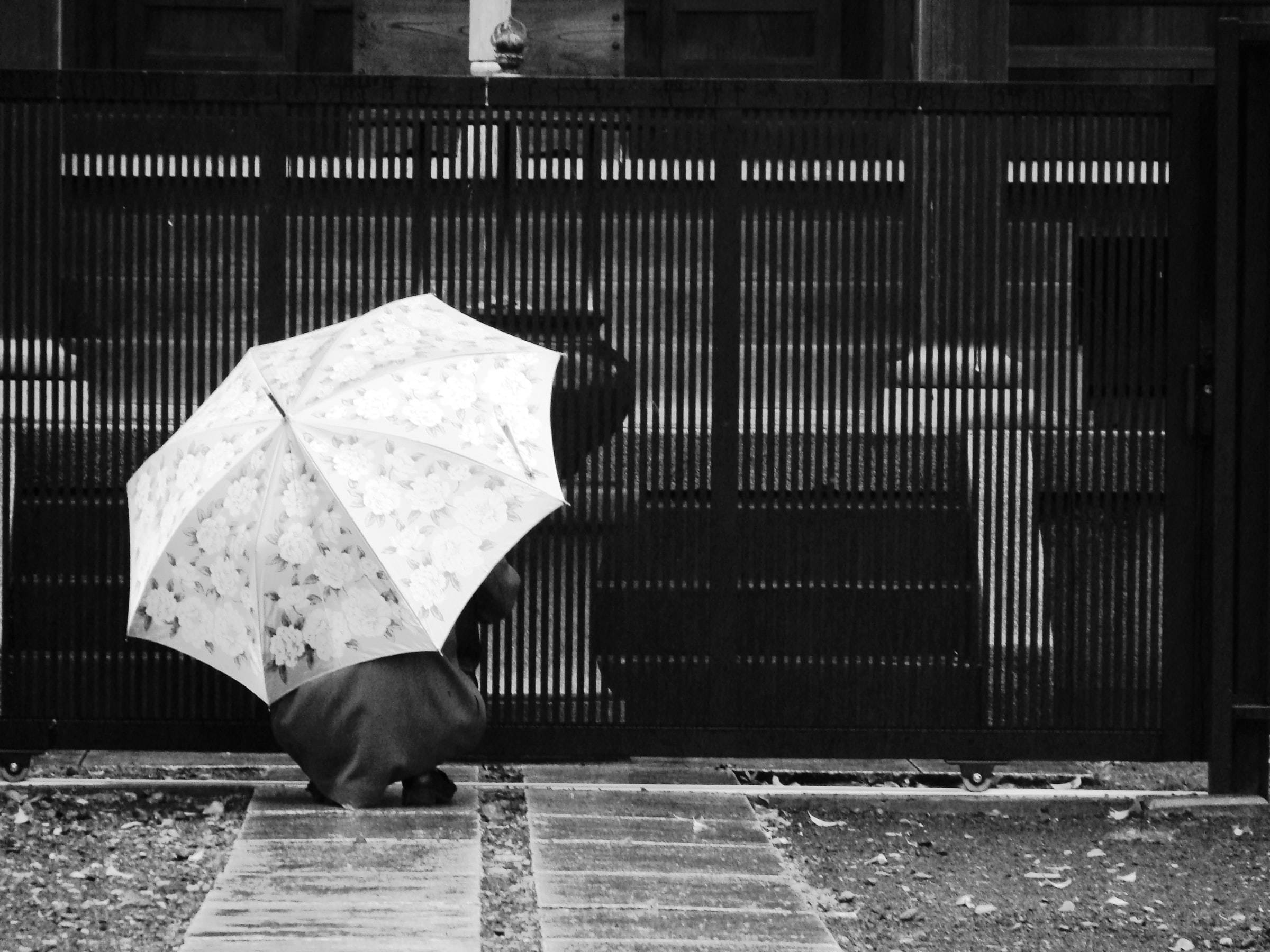 Remembrance 桌布 壁紙 — 一個人蹲在花紋雨傘下面，在濕石路旁邊，前面是深色木門。