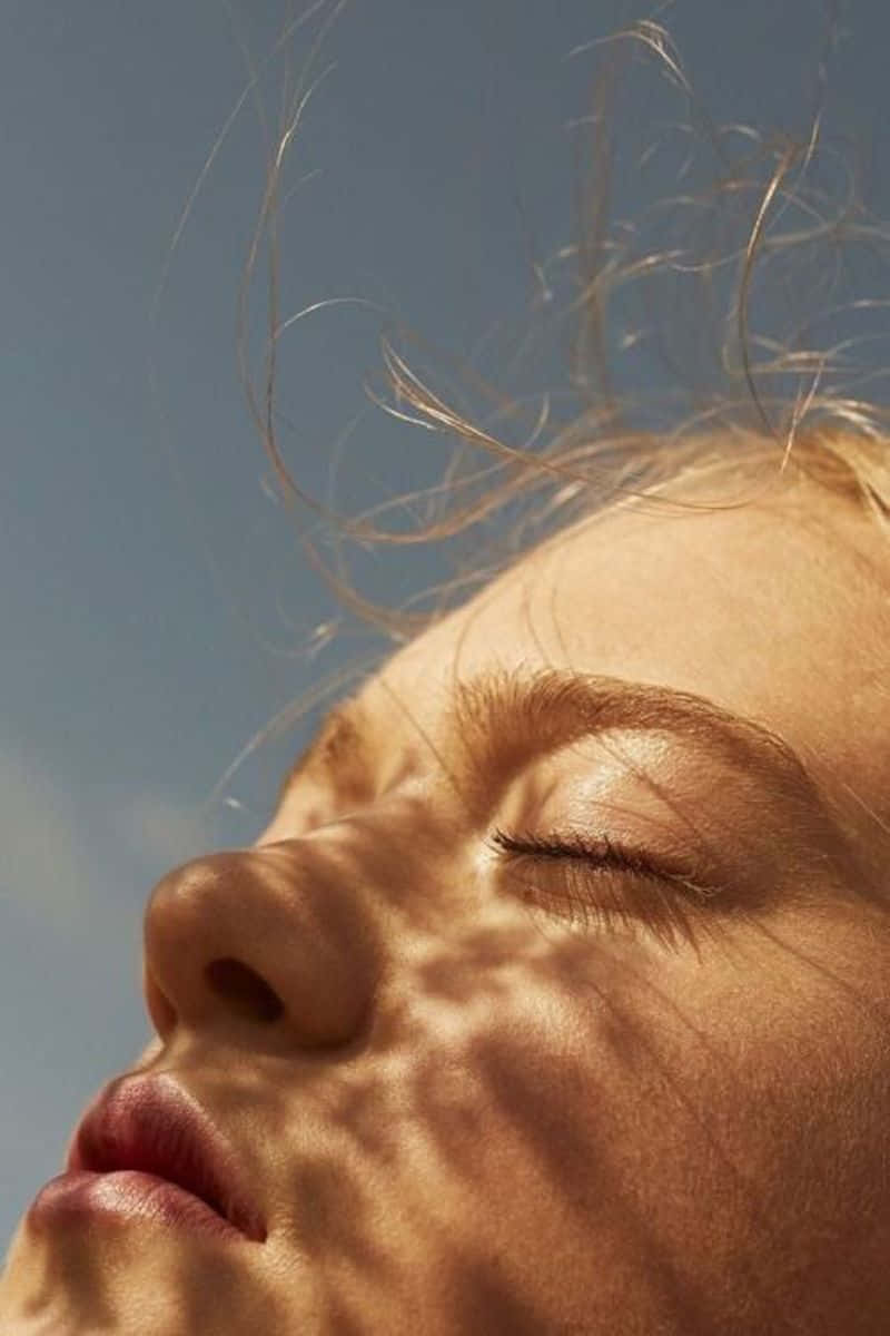 emotion 桌布 壁紙 — 一個人閉上眼睛的臉部特寫，沐浴在溫暖的陽光中，陽光在皮膚上投下樹葉的影子，背景是清澈的藍天。