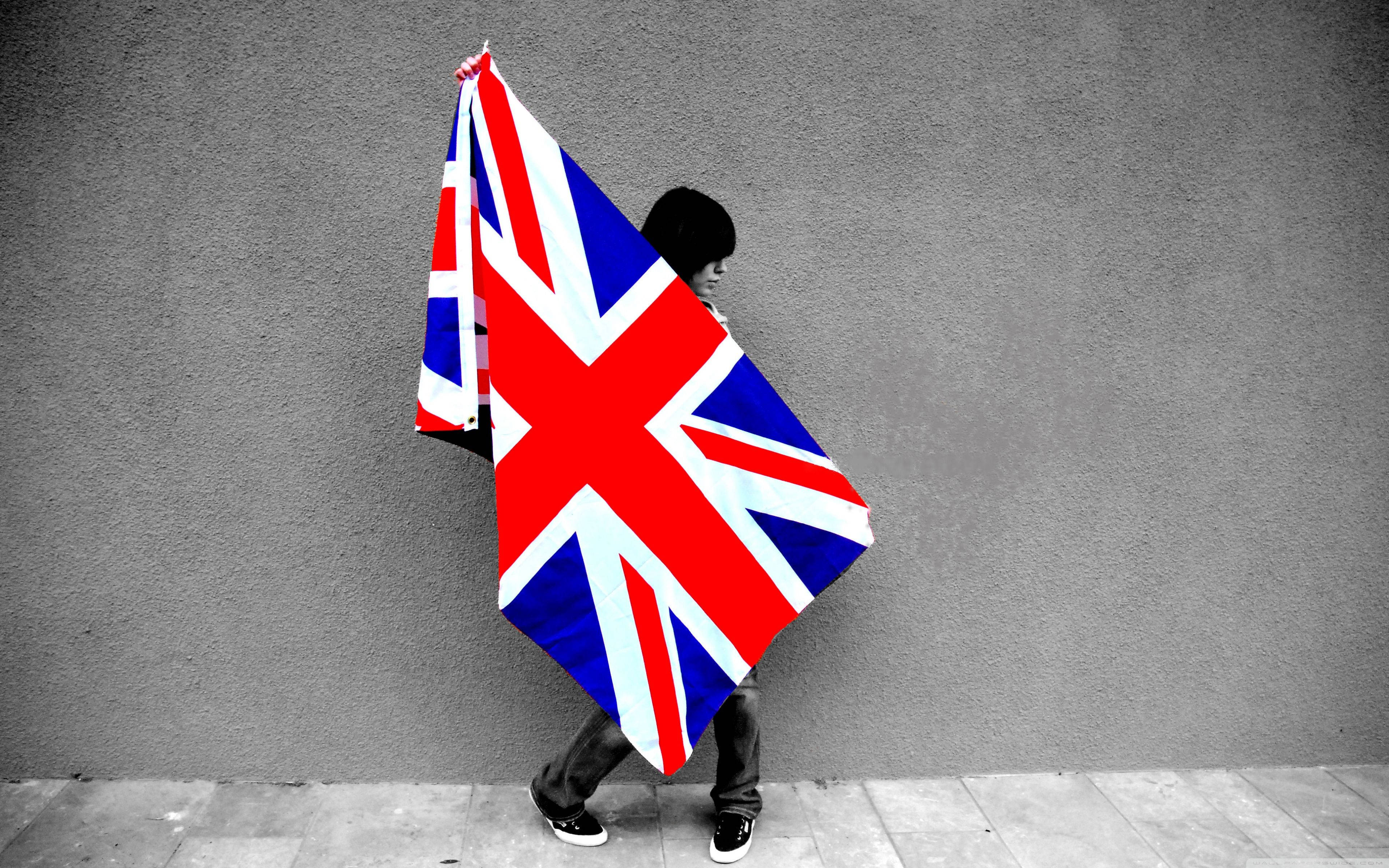 United Kingdom Flag 桌布 壁紙 — 一個人靠著灰色牆壁，手持一面鮮豔的英國國旗，旗幟以選擇性色彩遮蓋了他們的臉龐。