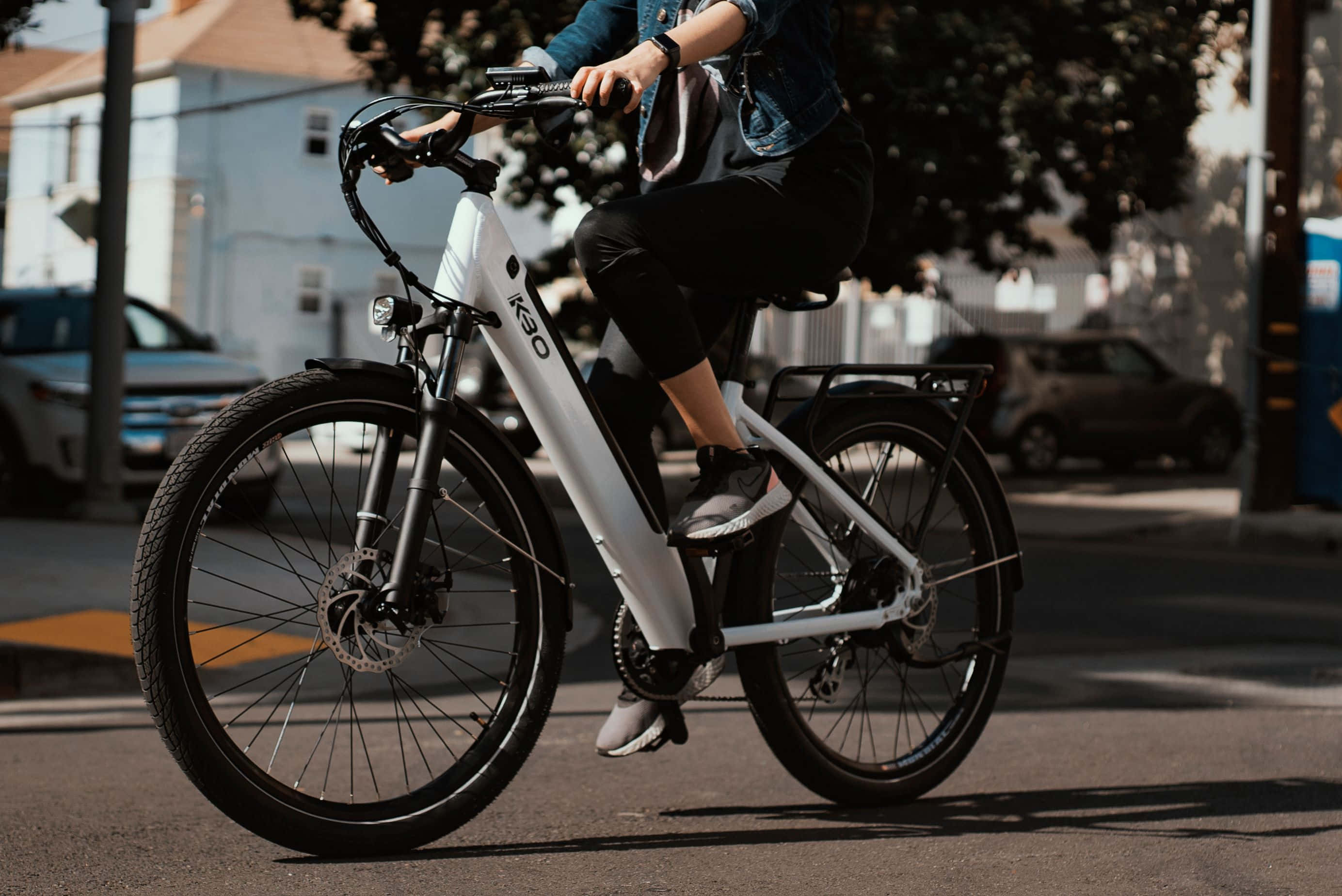 Electric Bike 桌布 壁紙 — 一個人騎著白色電動腳踏車，沿著陽光明媚的城市街道行駛，背景有停泊的汽車和樹木。