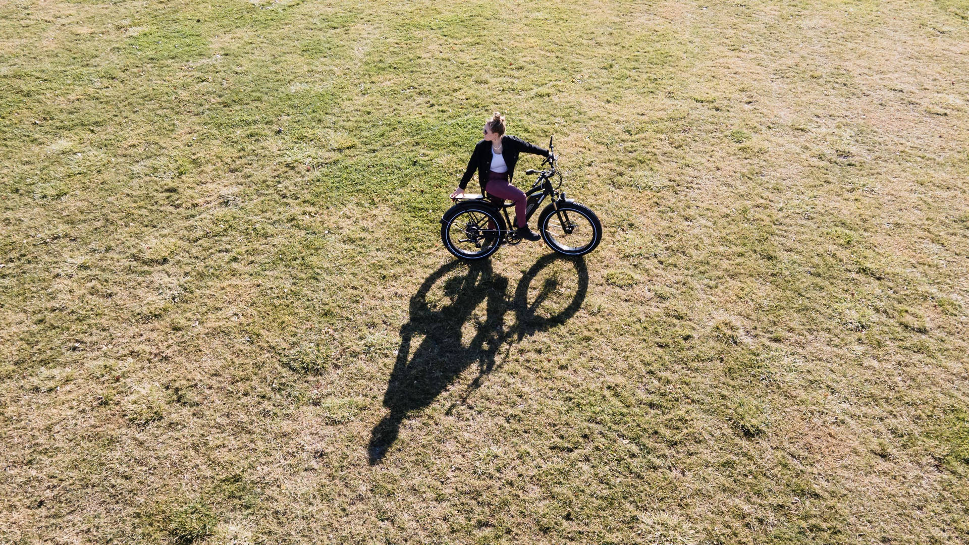 bike riding 桌布 壁紙 — 一個人騎著黑色電動腳踏車，在廣闊的乾草地田野上，在明亮的陽光下投下長長的影子。