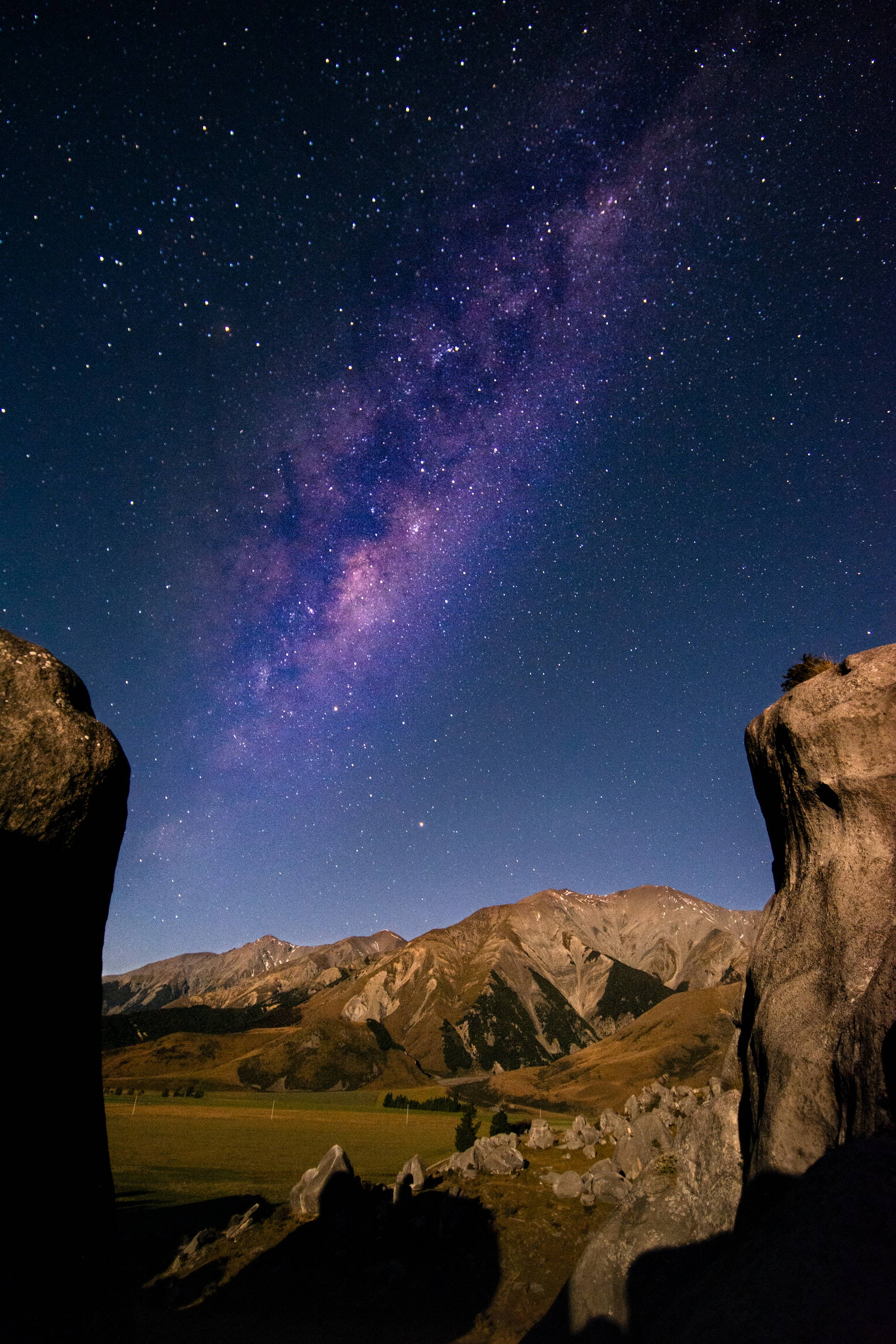 夜空 桌布 壁紙 — 一個令人嘆為觀止的夜空，佈滿星星和銀河，拱形橫跨崎嶇的山脈和一個由黑暗岩石框住的草地山谷。