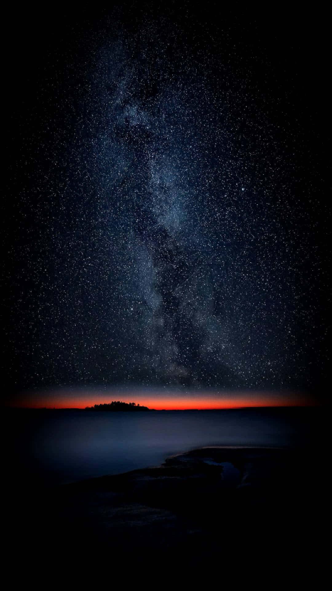 Calming iPhone 桌布 壁紙 — 一個令人嘆為觀止的夜空，佈滿星星和銀河，拱形在一個黑暗的岩石海岸線剪影之上，帶有發光的橙色地平線。