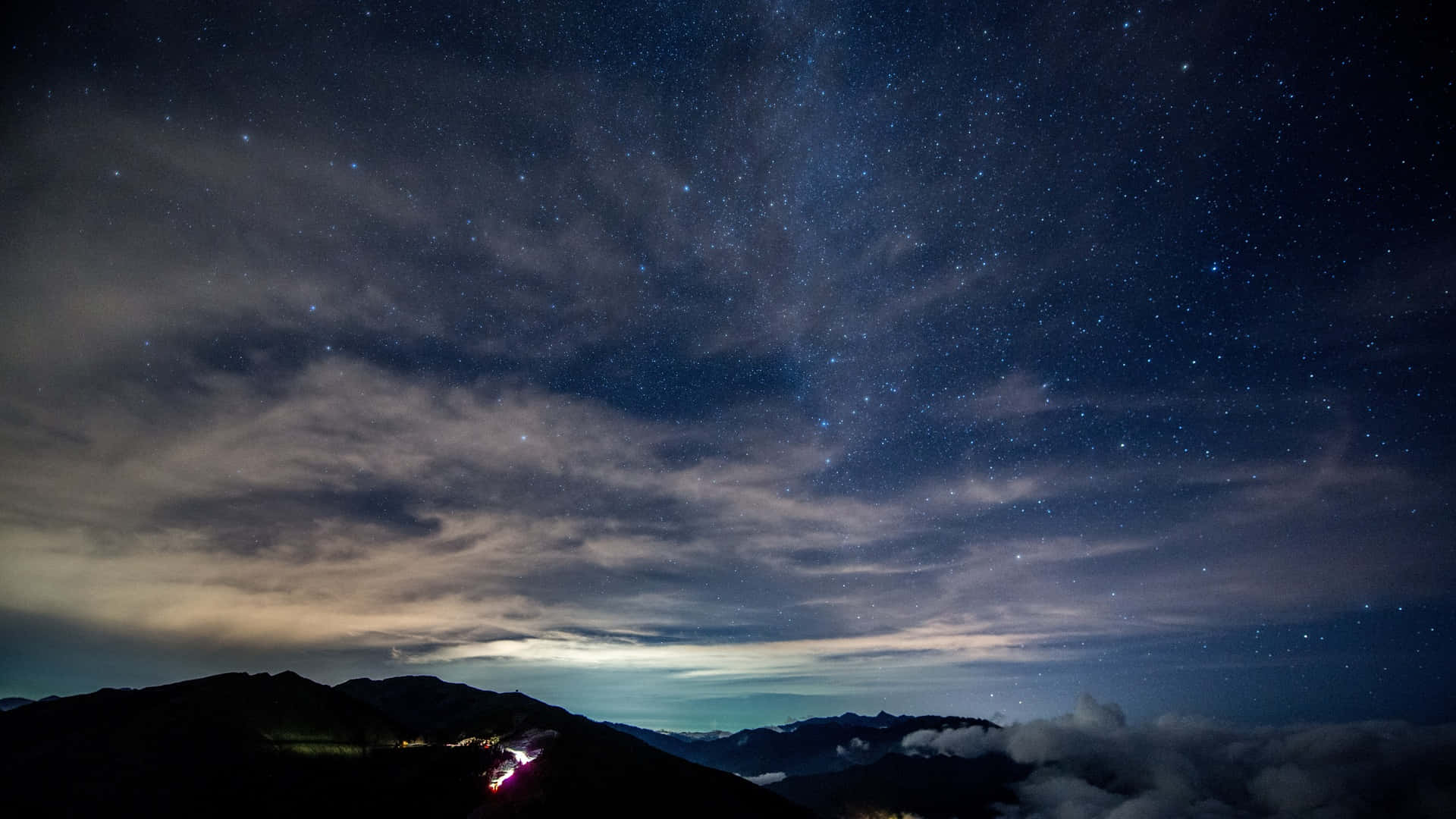 Dark Sky 桌布 壁紙 — 一個令人嘆為觀止的夜空，佈滿星星和縷縷雲彩，俯瞰著山脊，遠處的燈光蜿蜒穿過山谷。