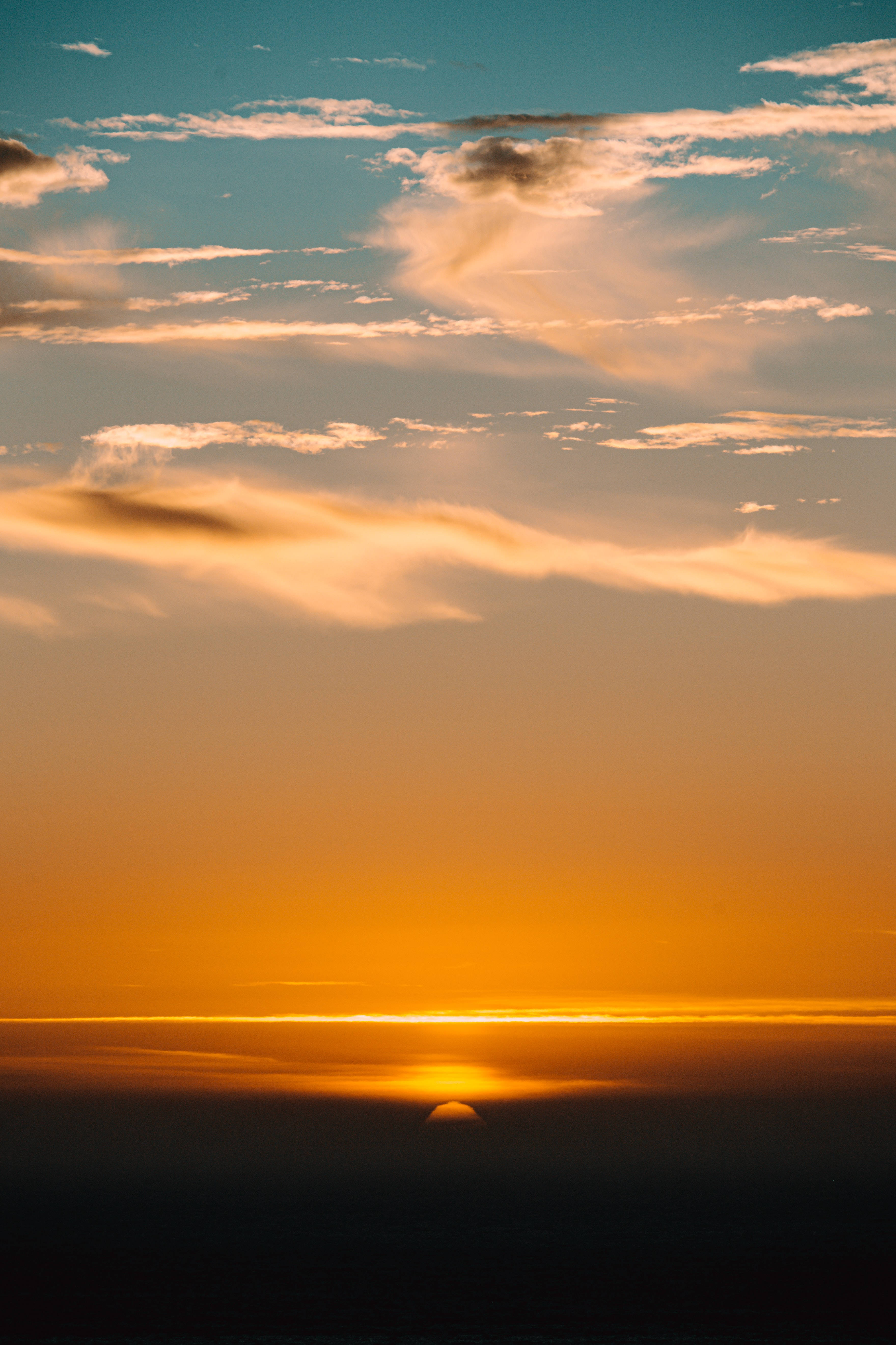 Beautiful Sky 桌布 壁紙 — 一個令人嘆為觀止的日落，發光的橙色太陽沉入黑暗的地平線下，將溫暖的光線投射在散落的雲朵上，映襯著青綠色的天空。