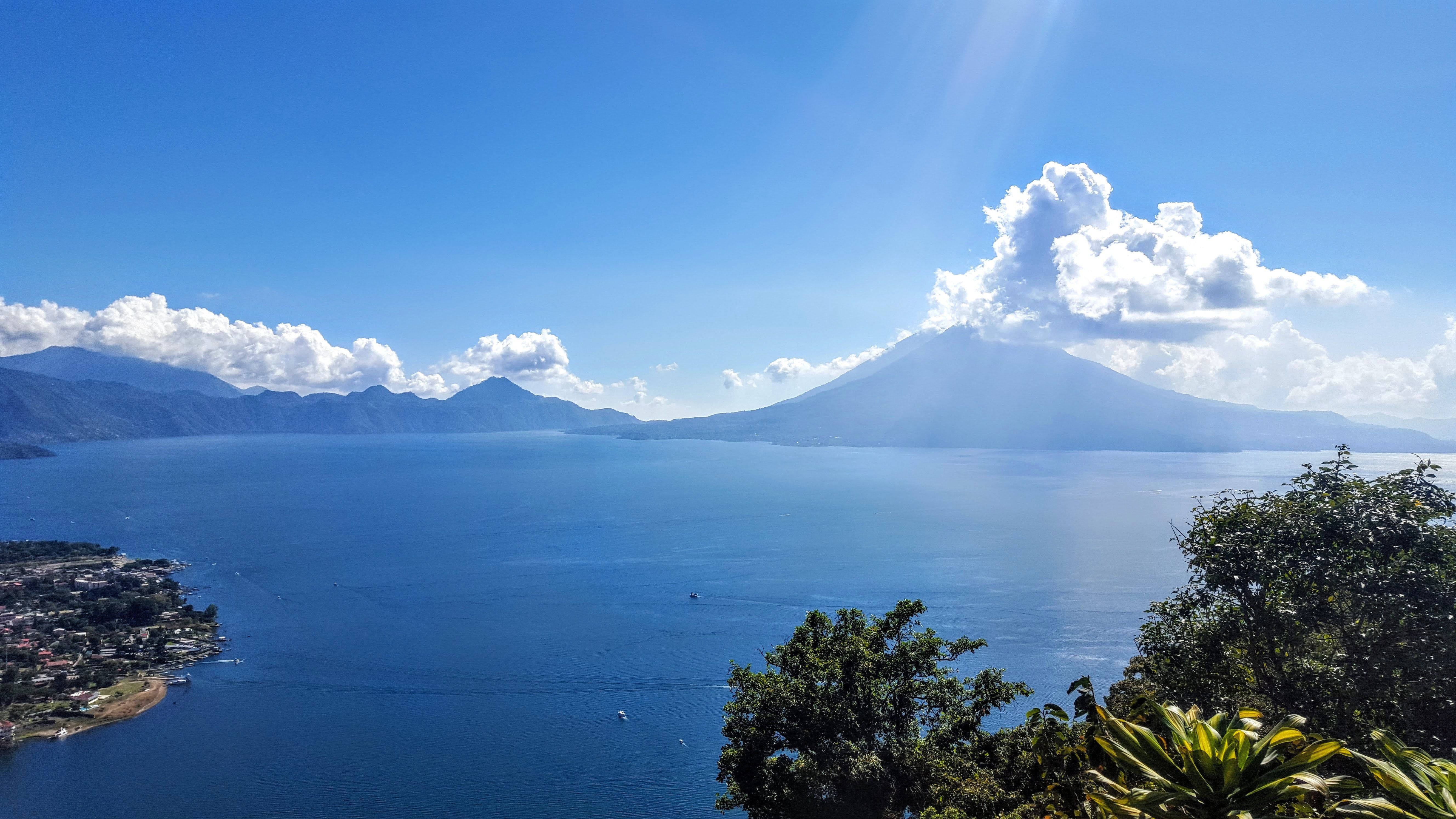 Guatemala 桌布 壁紙 — 一個令人嘆為觀止的景色，一座火山從廣闊的藍色湖泊中升起，在明亮的陽光天空下，散佈著雲朵。