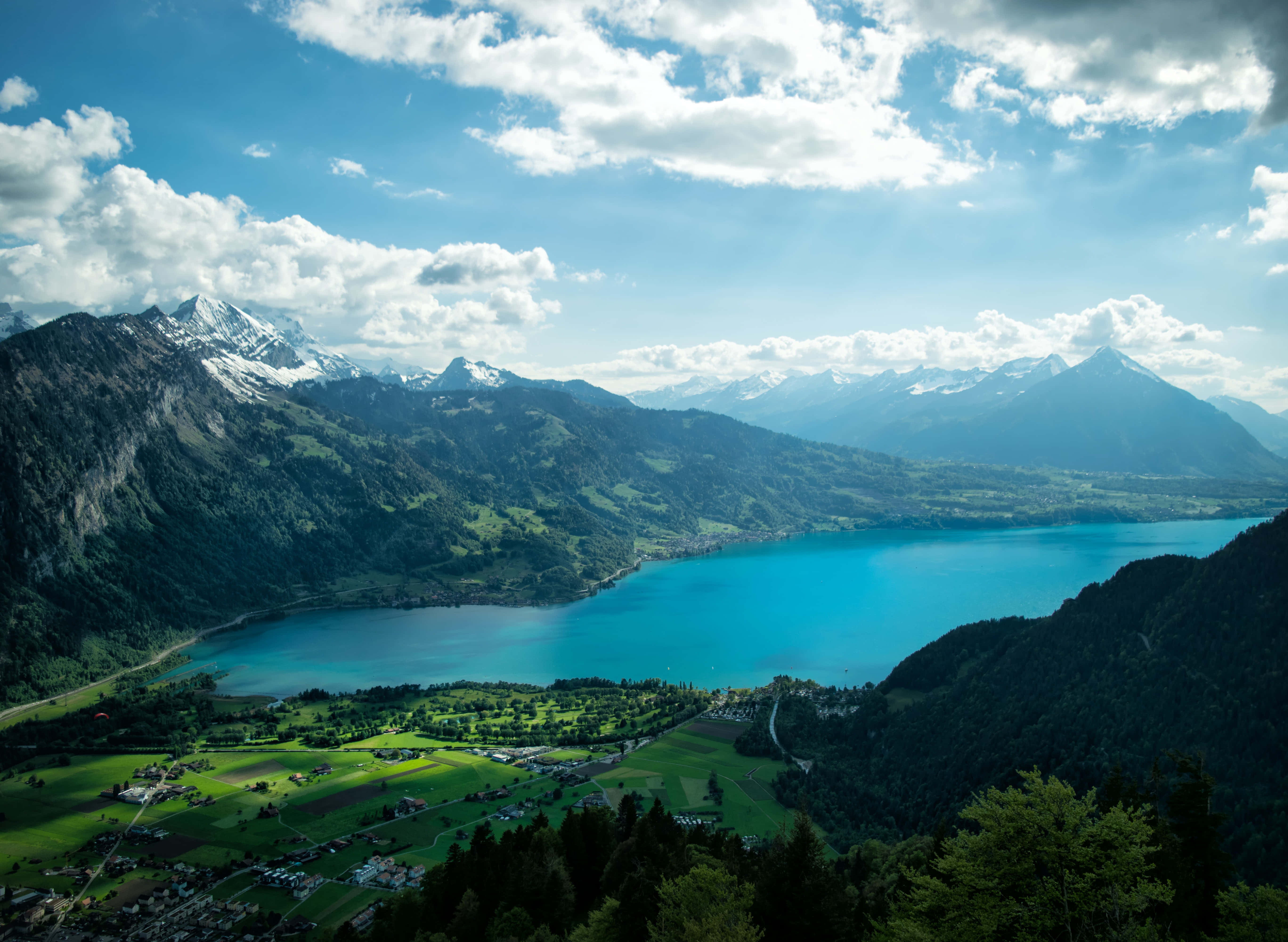 Beautiful Mountain Lake 桌布 壁紙 — 一個令人嘆為觀止的高山湖泊，湖水呈藍綠色，坐落在白雪覆蓋的山峰和翠綠的山谷之間，上方是明亮的藍天和蓬鬆的白雲。