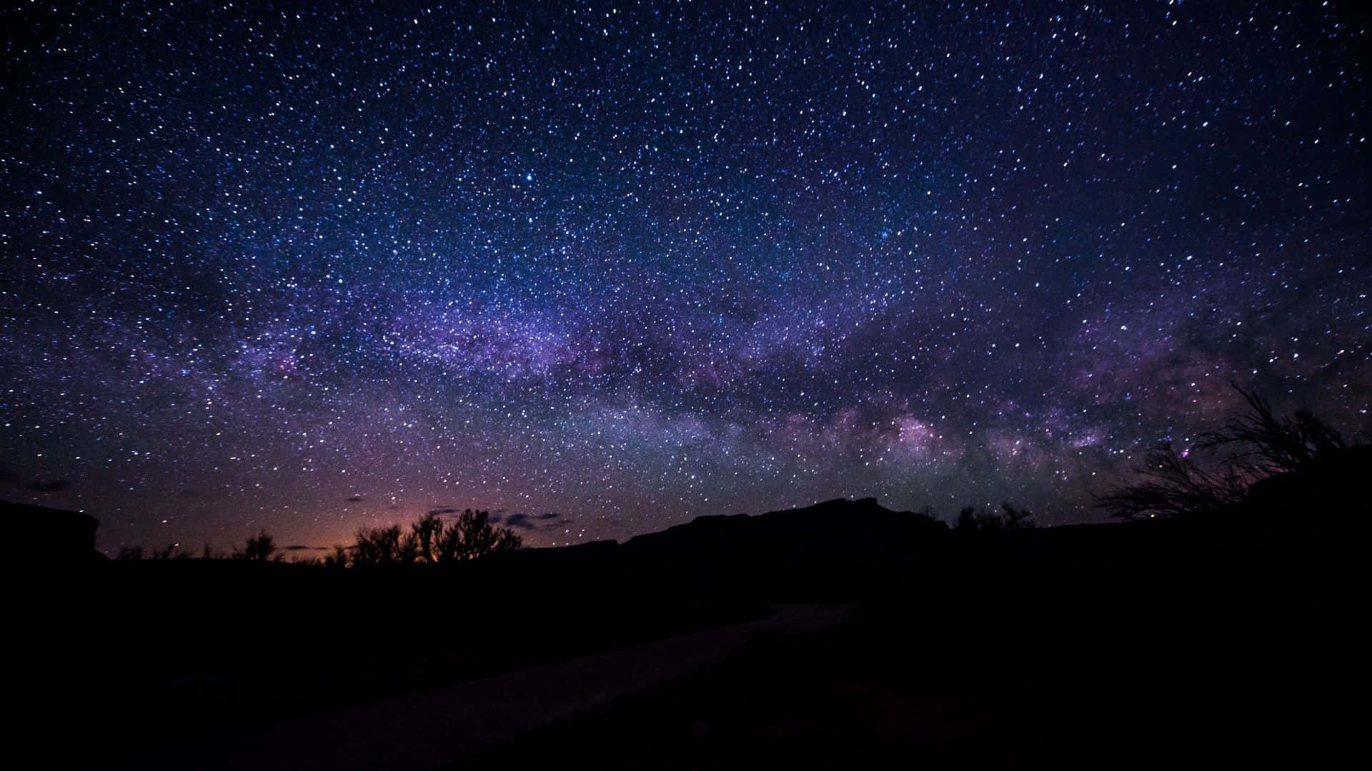 黑夜 桌布 壁紙 — 一個令人屏息的夜空，充滿無數星星和一條充滿活力的紫色銀河，拱在黑暗的山丘剪影之上。