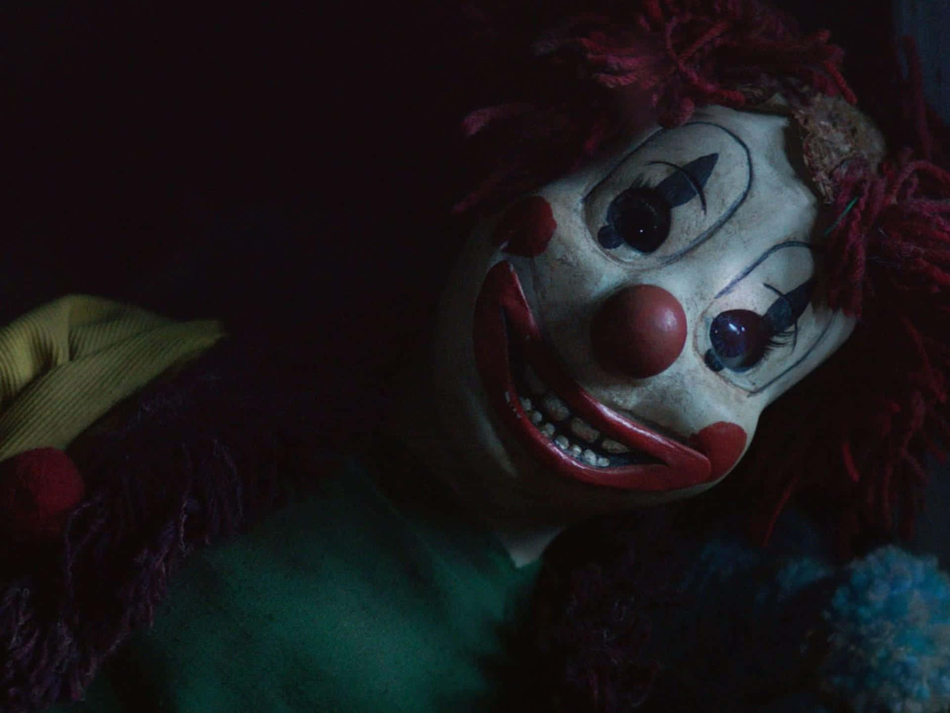 Cool Scary 桌布 壁紙 — 一個令人毛骨悚然的小丑娃娃特寫，紅髮，在昏暗燈光下露出寬闊的牙齒笑容。