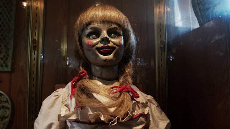 Annabelle 桌布 壁紙 — 一個令人毛骨悚然的復古娃娃，臉部有裂紋的瓷面和紅色絲帶，展示在昏暗木箱的玻璃後面。