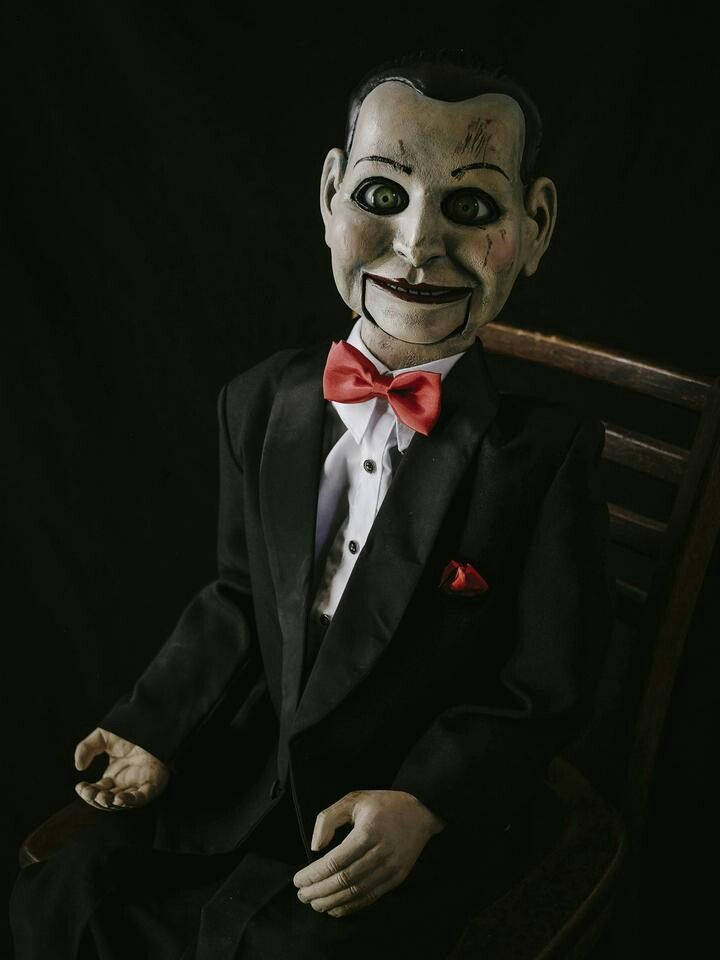 horror villains 桌布 壁紙 — 一個令人毛骨悚然的復古木偶，臉上塗有顏料，戴著紅色領結，坐在黑色背景前的木椅上。