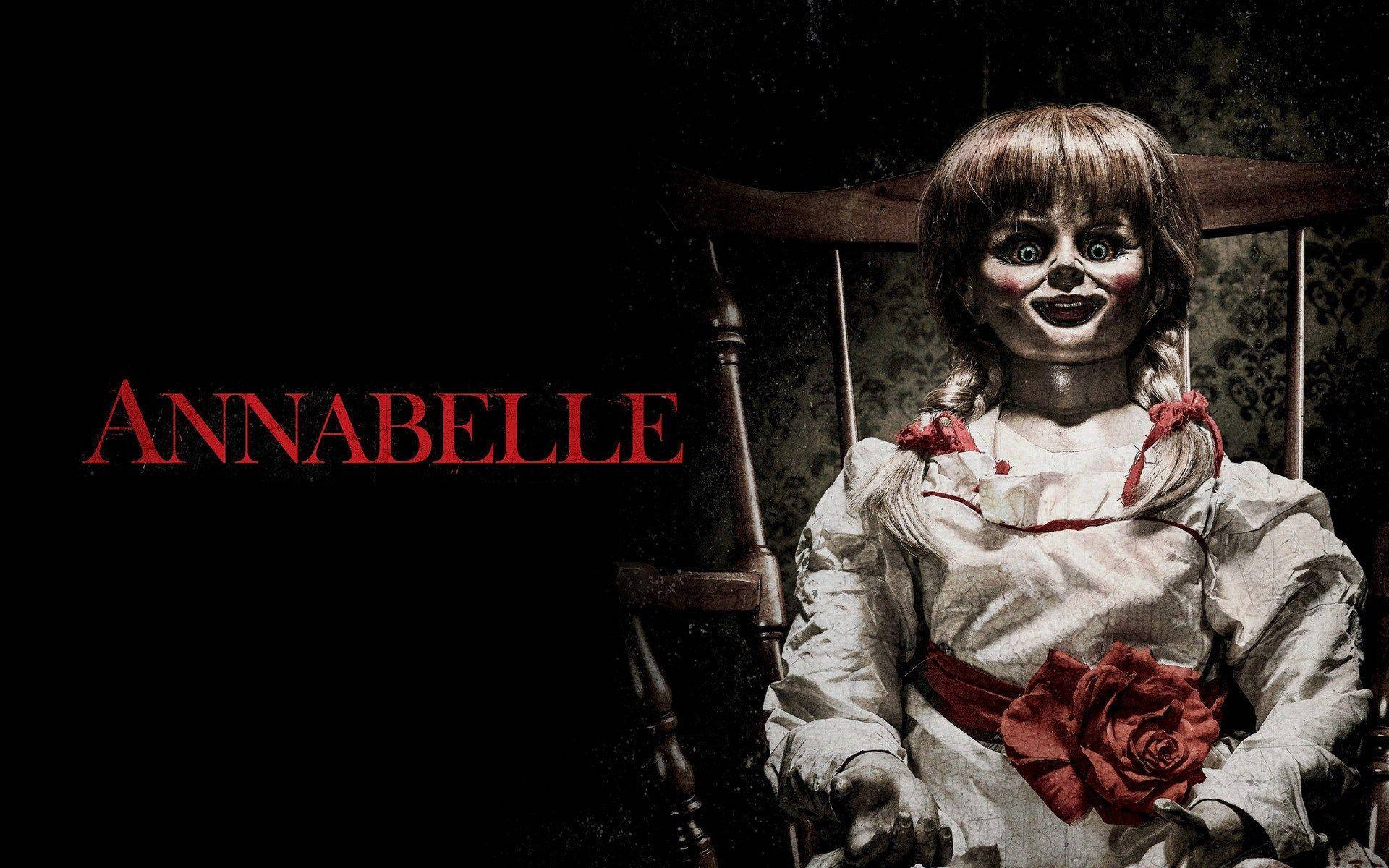 Annabelle 桌布 壁紙 — 一個令人毛骨悚然的洋娃娃，大眼睛，手持紅玫瑰，坐在木椅上，背景是深色壁紙牆壁，紅色標題為Annabelle。