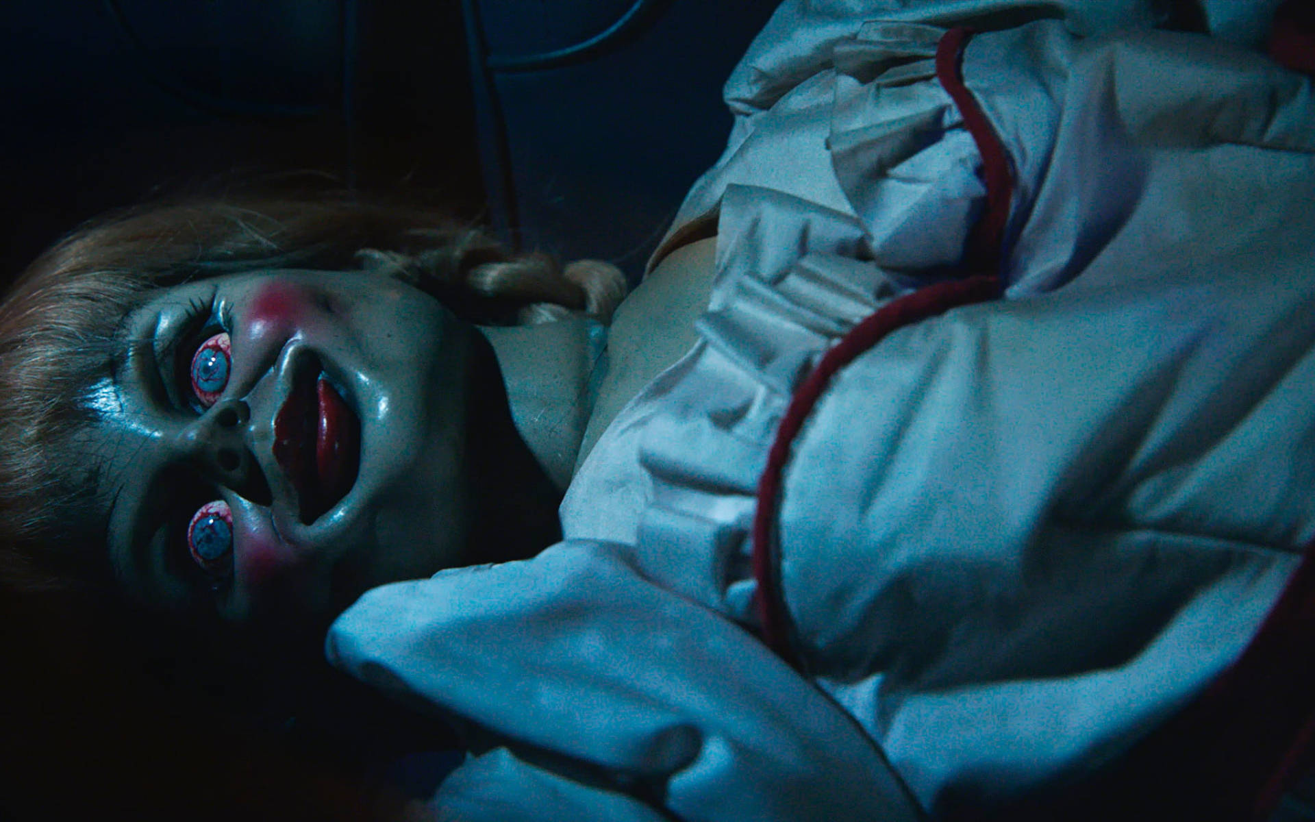 Annabelle 桌布 壁紙 — 一個令人毛骨悚然的洋娃娃，有著寬大發光的眼睛和紅唇，躺在昏暗的藍光中，營造出令人不寒而慄的恐怖氣氛。