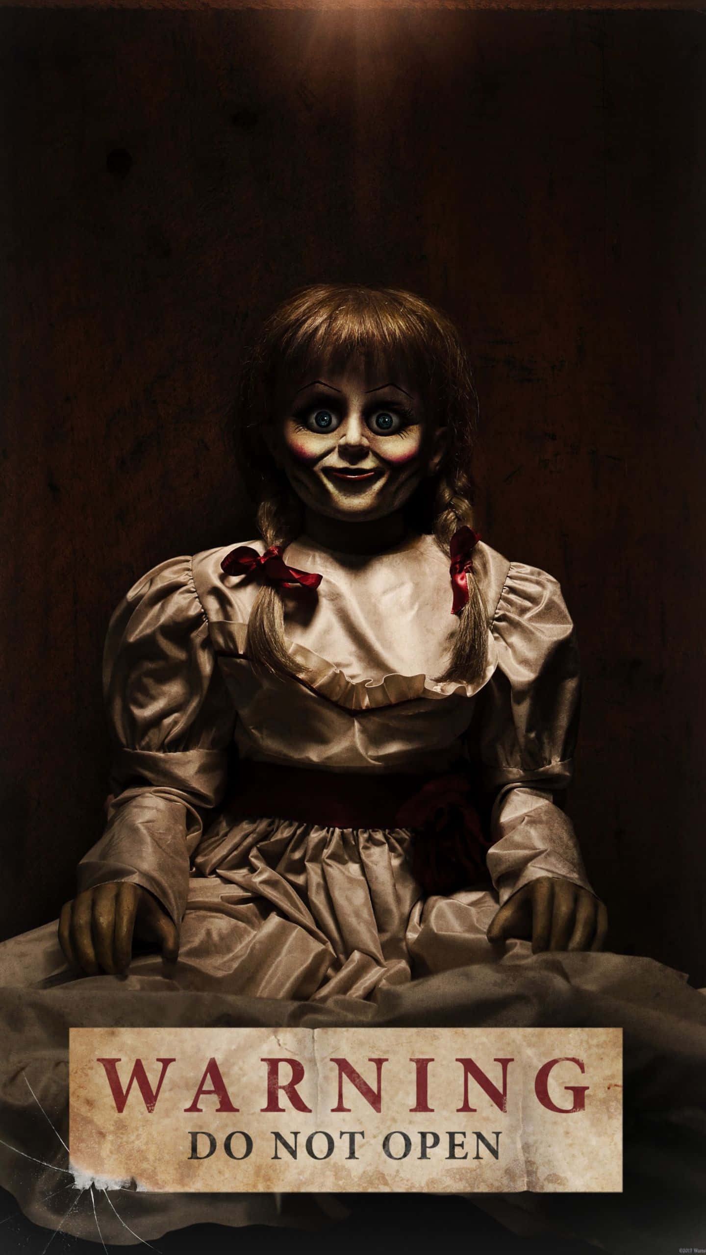 Annabelle 桌布 壁紙 — 一個令人毛骨悚然的洋娃娃，睜大眼睛，露出邪惡的笑容，坐在昏暗的燈光下，下面有一個警告標誌。
