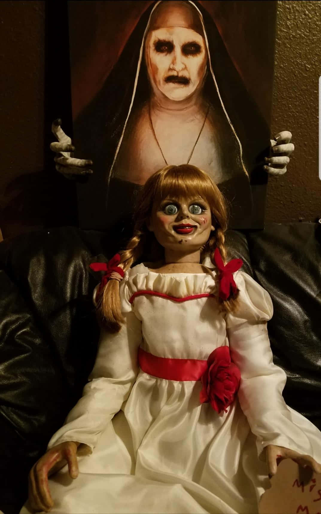 Annabelle 桌布 壁紙 — 一個令人毛骨悚然的洋娃娃，辮子頭髮上繫著紅絲帶，坐在黑色沙發上，沙發上方掛著一幅臉色蒼白、雙手如骷髏的修女畫像。