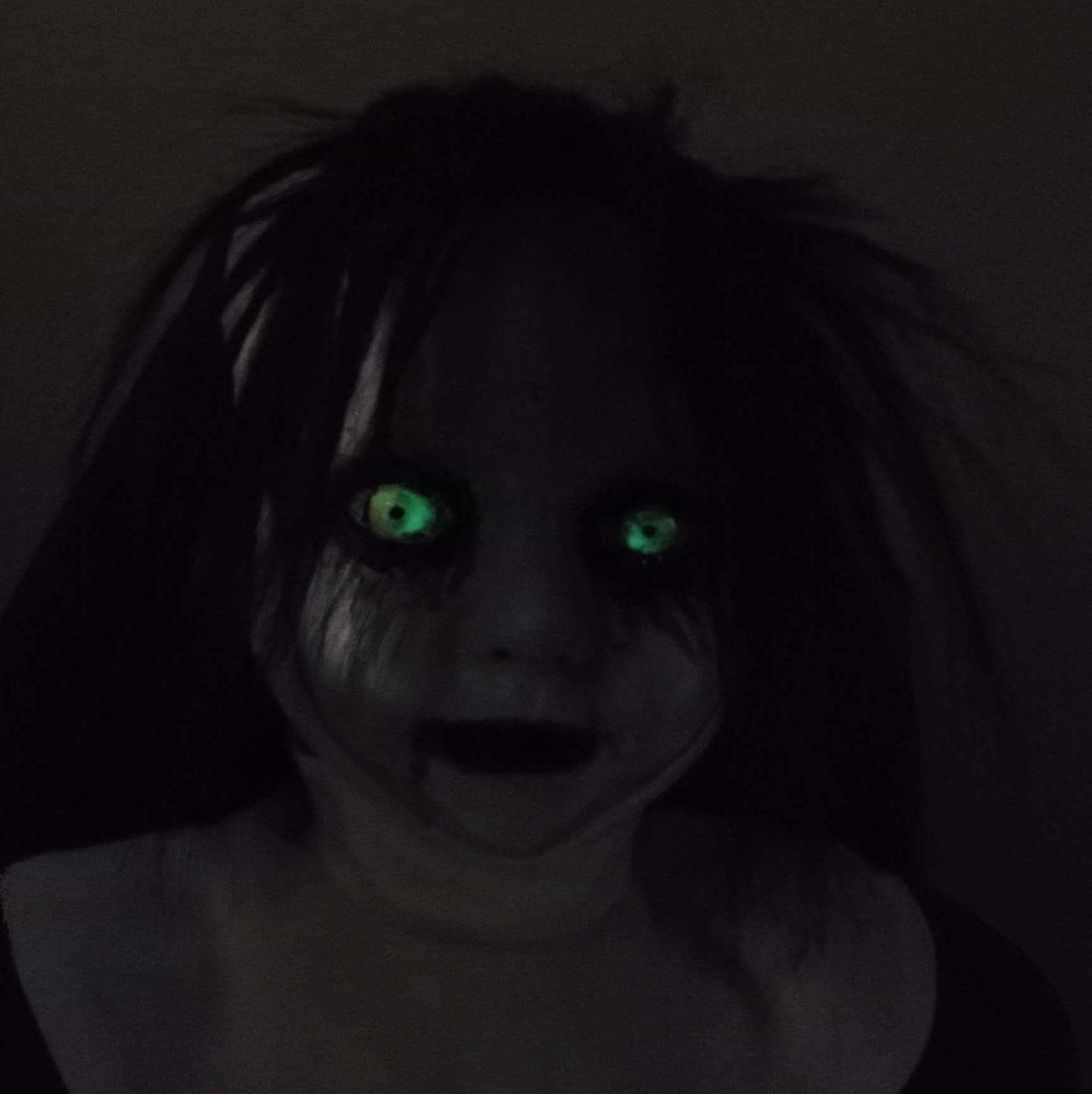 Creepy Halloween 桌布 壁紙 — 一個令人毛骨悚然的特寫，一個在近乎黑暗中擁有發光綠色眼睛和凌亂深色頭髮的娃娃。