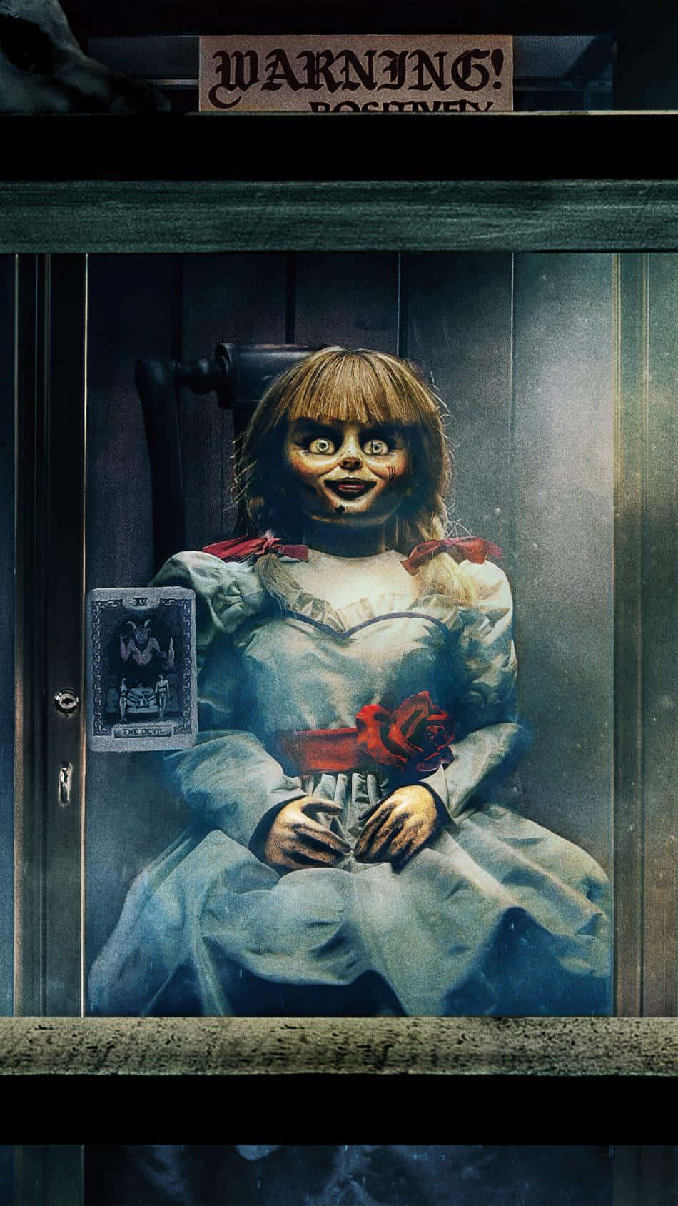 Annabelle 桌布 壁紙 — 一個令人毛骨悚然的瓷臉娃娃，大眼睛，手持紅玫瑰，坐在玻璃櫃中，上方有「警告！」標誌。