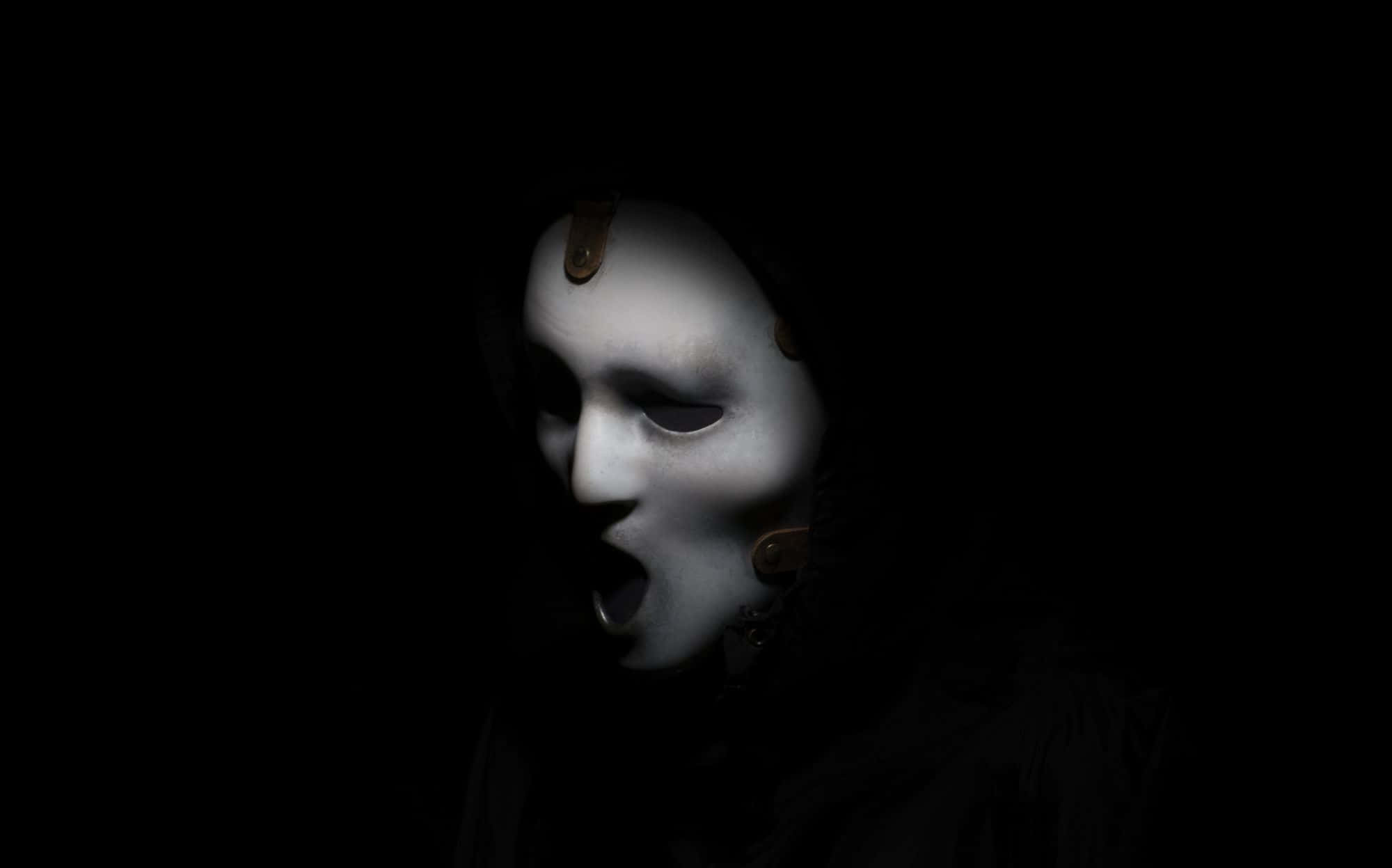 Ghost Face 桌布 壁紙 — 一個令人毛骨悚然的白色面具人物張開嘴巴從黑暗中出現，近距離特寫。