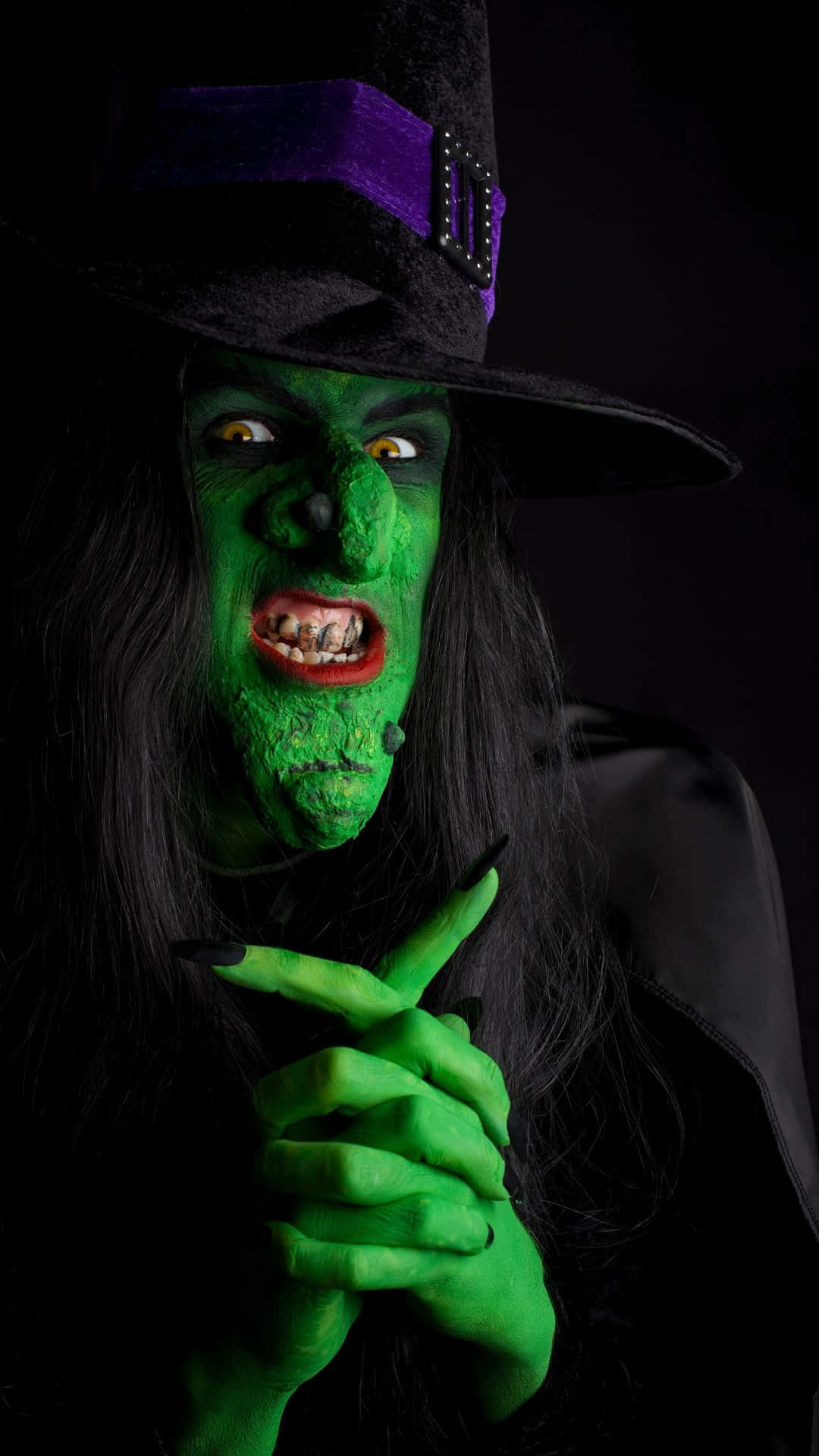 Horror Costumes 桌布 壁紙 — 一個令人畏懼的綠皮膚女巫，有著發光的黃色眼睛和黑色帽子，在黑暗背景下指責地指著。