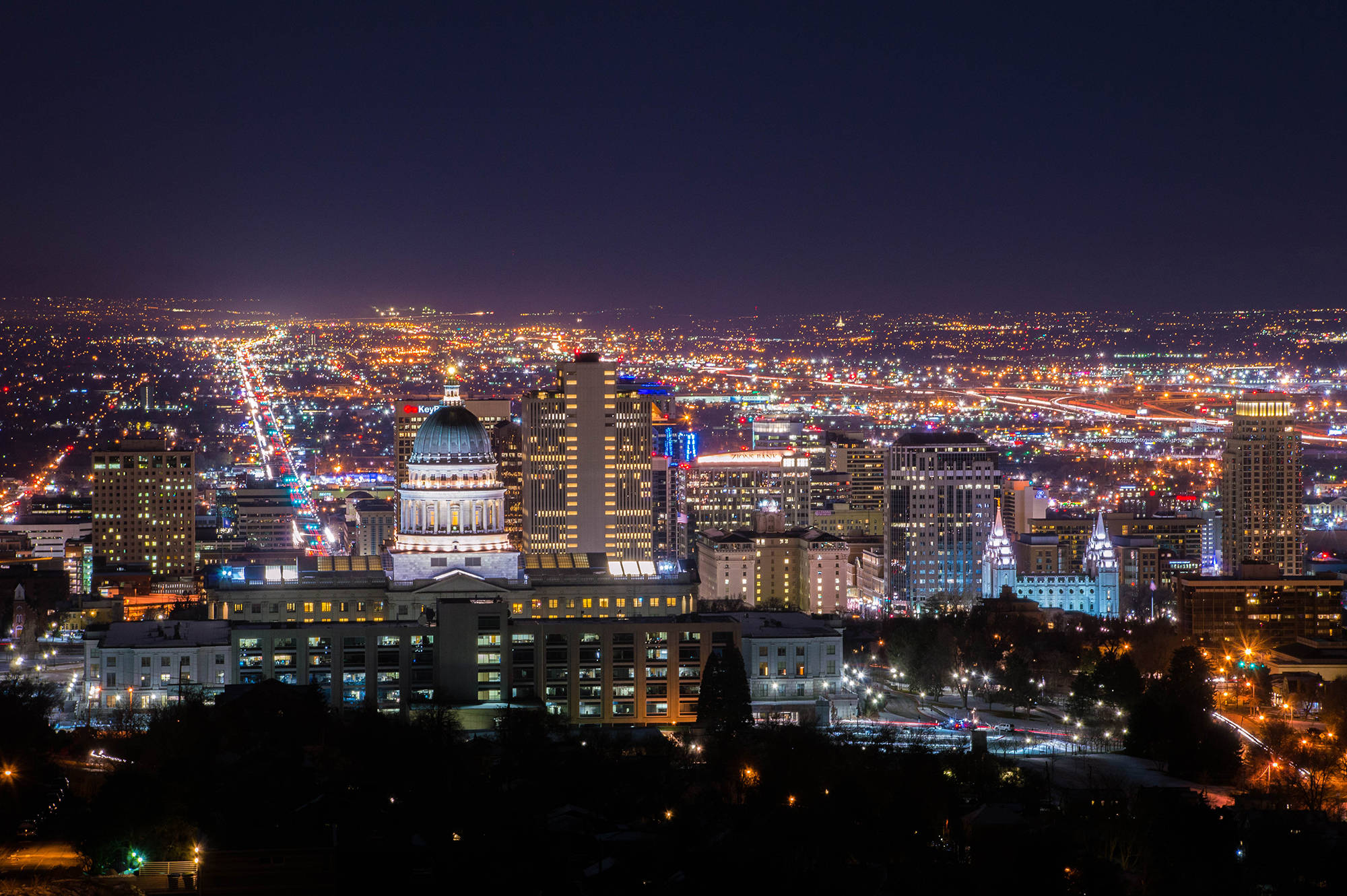 Salt Lake City 桌布 壁紙 — 一個令人目眩的夜景城市景觀，圓頂國會大廈在黑暗的天空下被照亮，周圍環繞著閃爍的摩天大樓和流動的交通燈光。