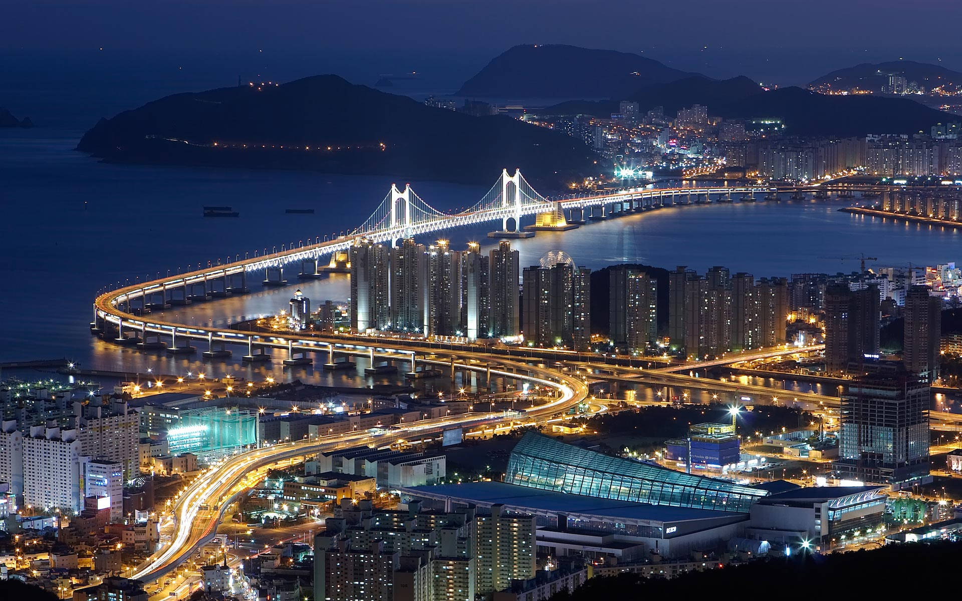 South Korea 桌布 壁紙 — 一個令人眼花撩亂的夜景，展現了廣闊的海濱城市，一座發光的吊橋橫跨被山丘環繞的黑暗海灣。