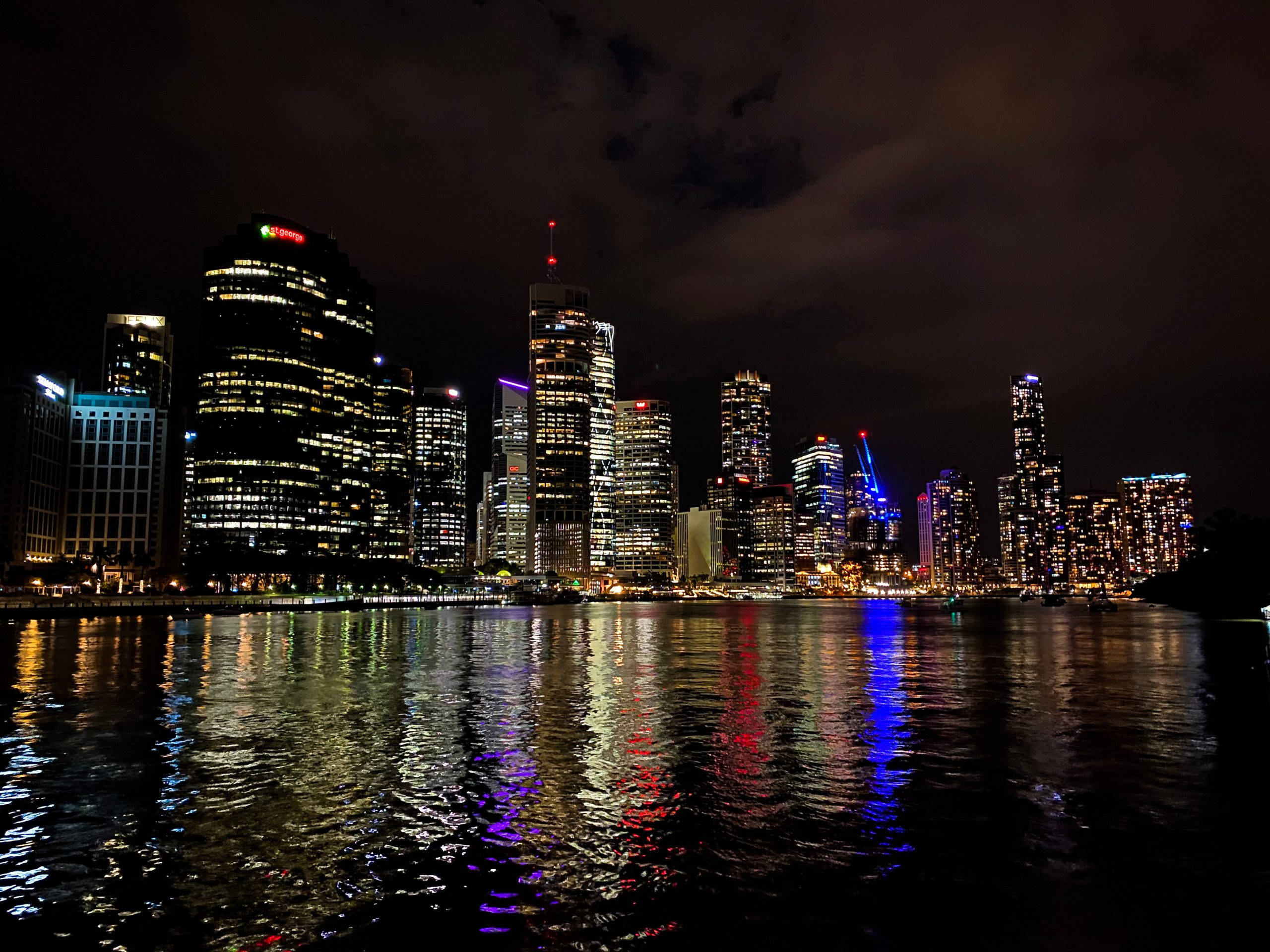 City At Night 桌布 壁紙 — 一個令人眼花撩亂的夜景城市景觀，摩天大樓在黑暗的天空中閃耀，它們的燈光在水面上閃爍。