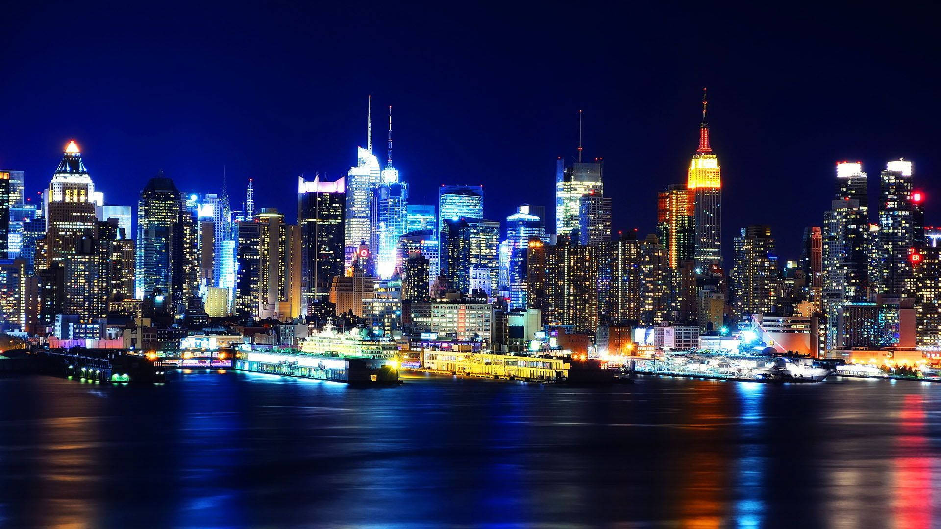 夜空 桌布 壁紙 — 一個令人眼花撩亂的夜景城市景觀，高聳的摩天大樓閃爍著霓虹燈光，倒映在黑暗的水道中。