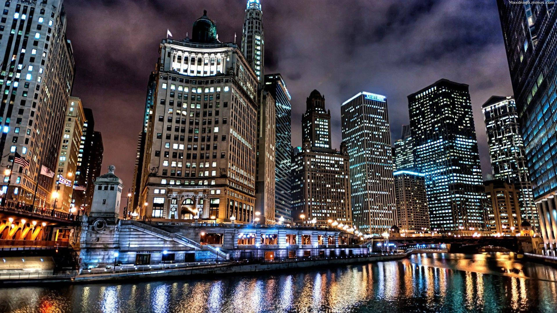 Skyscraper 桌布 壁紙 — 一個令人眼花撩亂的夜景城市景觀，高聳的摩天大樓在平靜的河面上反射出鮮豔的燈光，天空多雲。