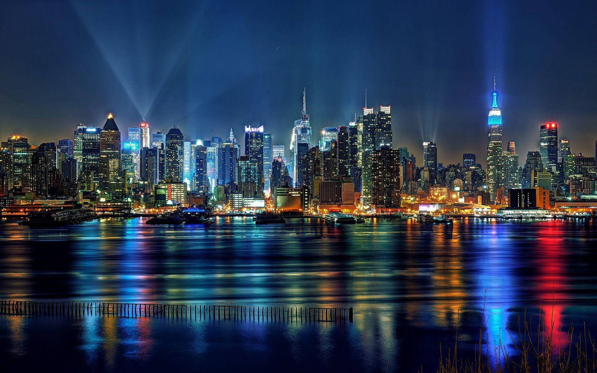New York 桌布 壁紙 — 一個令人眼花撩亂的夜景城市景觀，摩天大樓在光線照射下閃閃發光，倒映在黑暗的水面前景中。