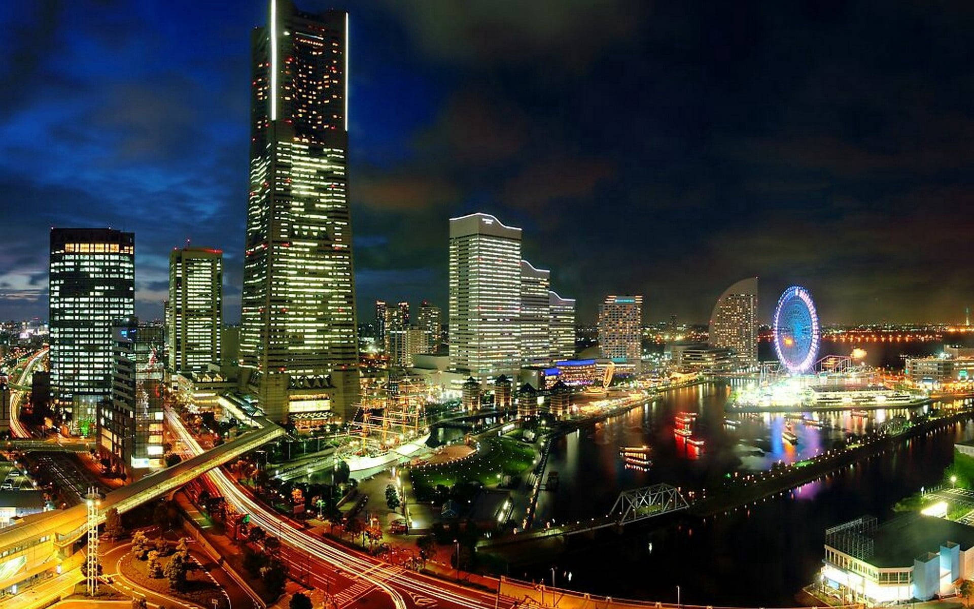 Yokohama 桌布 壁紙 — 一個令人眼花撩亂的夜間城市景觀，高聳的摩天大樓在黑暗的天空中閃耀，河流反射燈光，一個巨大的摩天輪照亮了海濱。