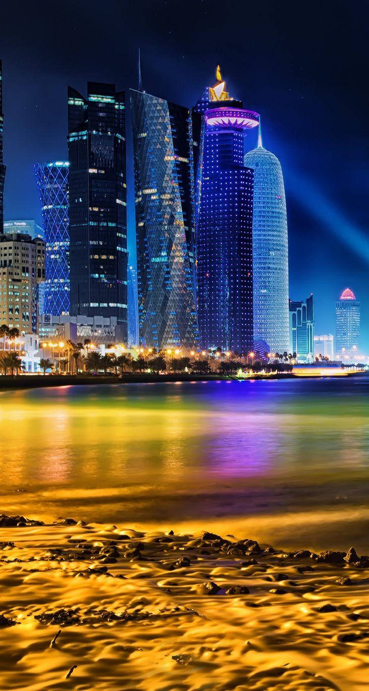 夜空 桌布 壁紙 — 一個令人眼花繚亂的夜景城市景觀，燈火通明的摩天大樓在平靜的水面上反射出鮮豔的色彩，旁邊是沙灘。