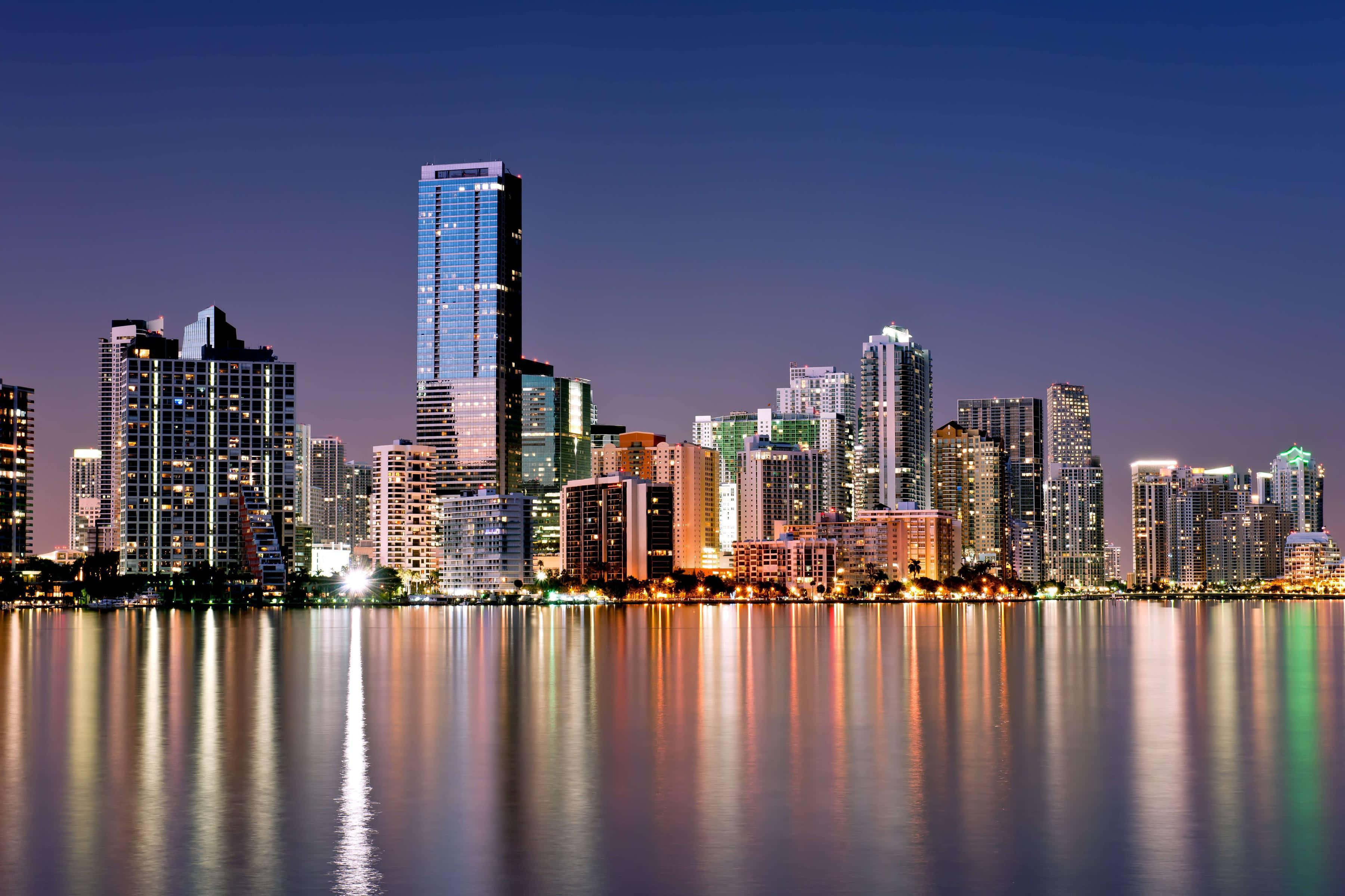 Miami 4K 桌布 壁紙 — 一個令人驚嘆的夜景城市景觀，摩天大樓在深藍色天空下，在平靜的水面上反射出鮮豔的燈光。