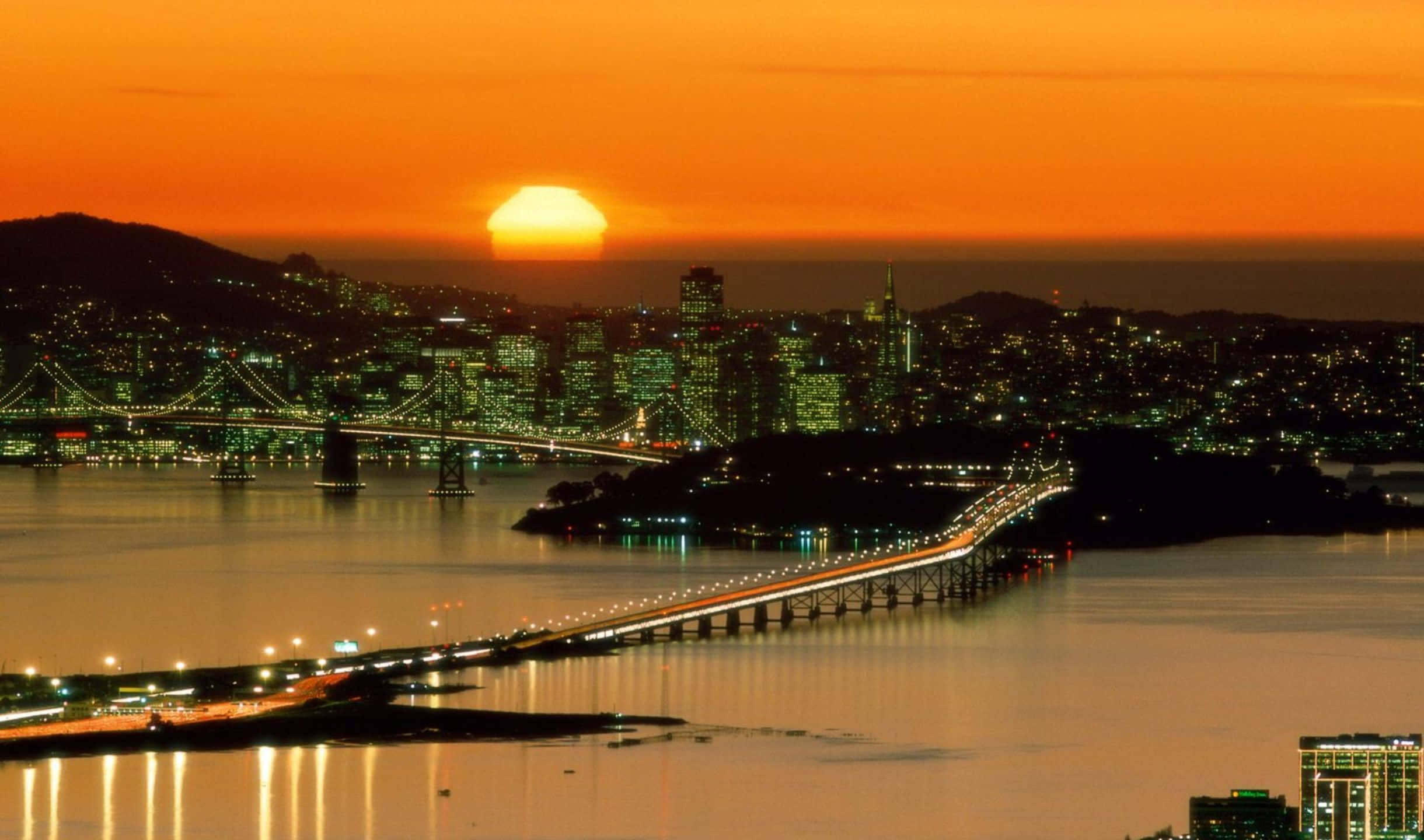 2440x1440 San Francisco 桌布 壁紙 — 一個令人驚嘆的日落，城市景觀中閃爍的橋樑和水面反射溫暖的橙色和綠色燈光。