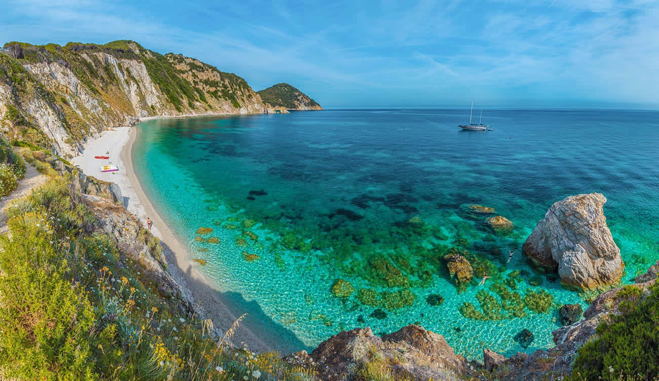 Italian Island 桌布 壁紙 — 一個令人驚嘆的海岸小海灣，海水呈碧綠色，清澈得可以看到下面的岩石，一艘孤獨的帆船在明亮的藍天下漂流。